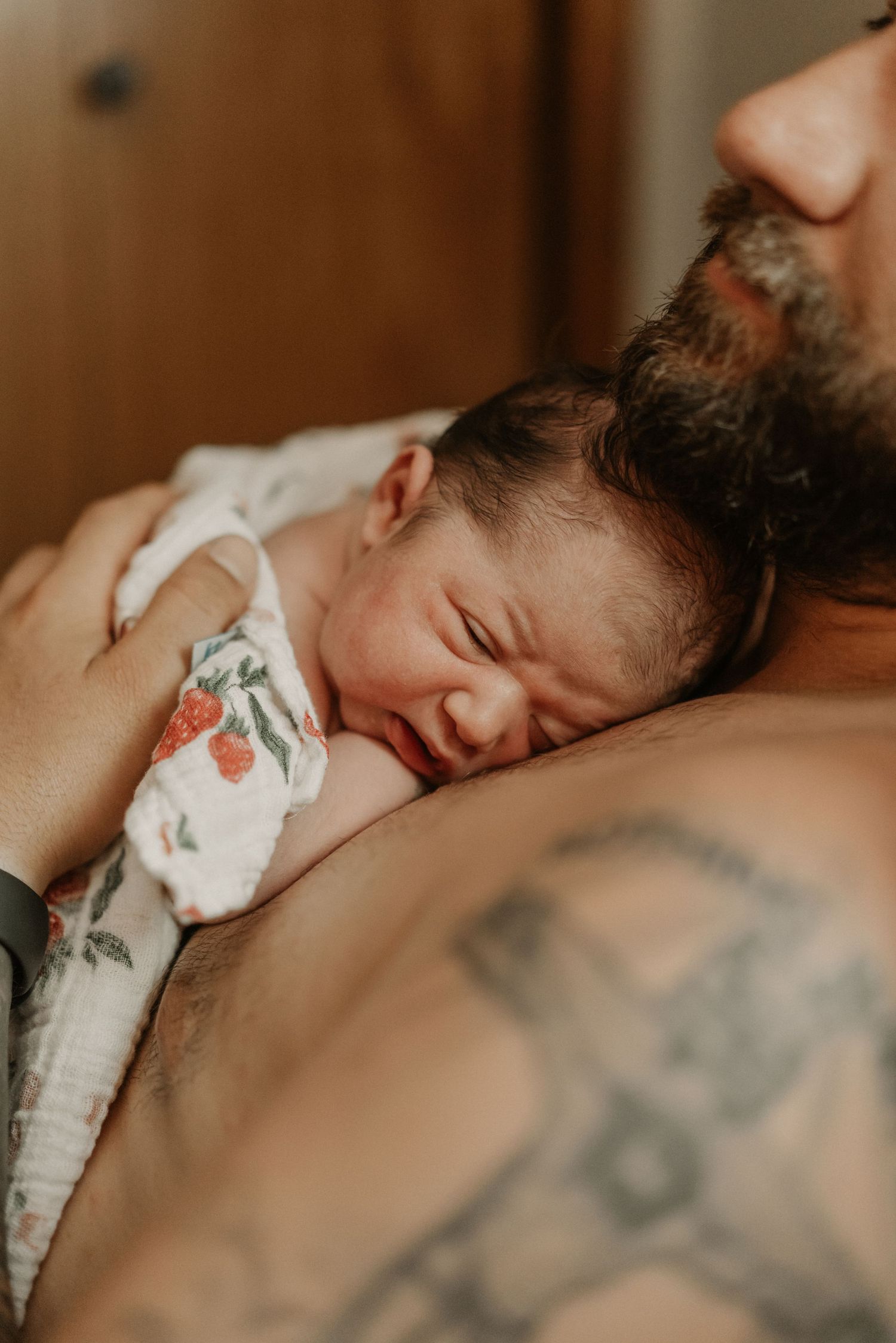Newborn baby sleeping peacefully on father's tattooed chest wrapped in a floral muslin swaddle blanket. Anchorage-birth-photographer