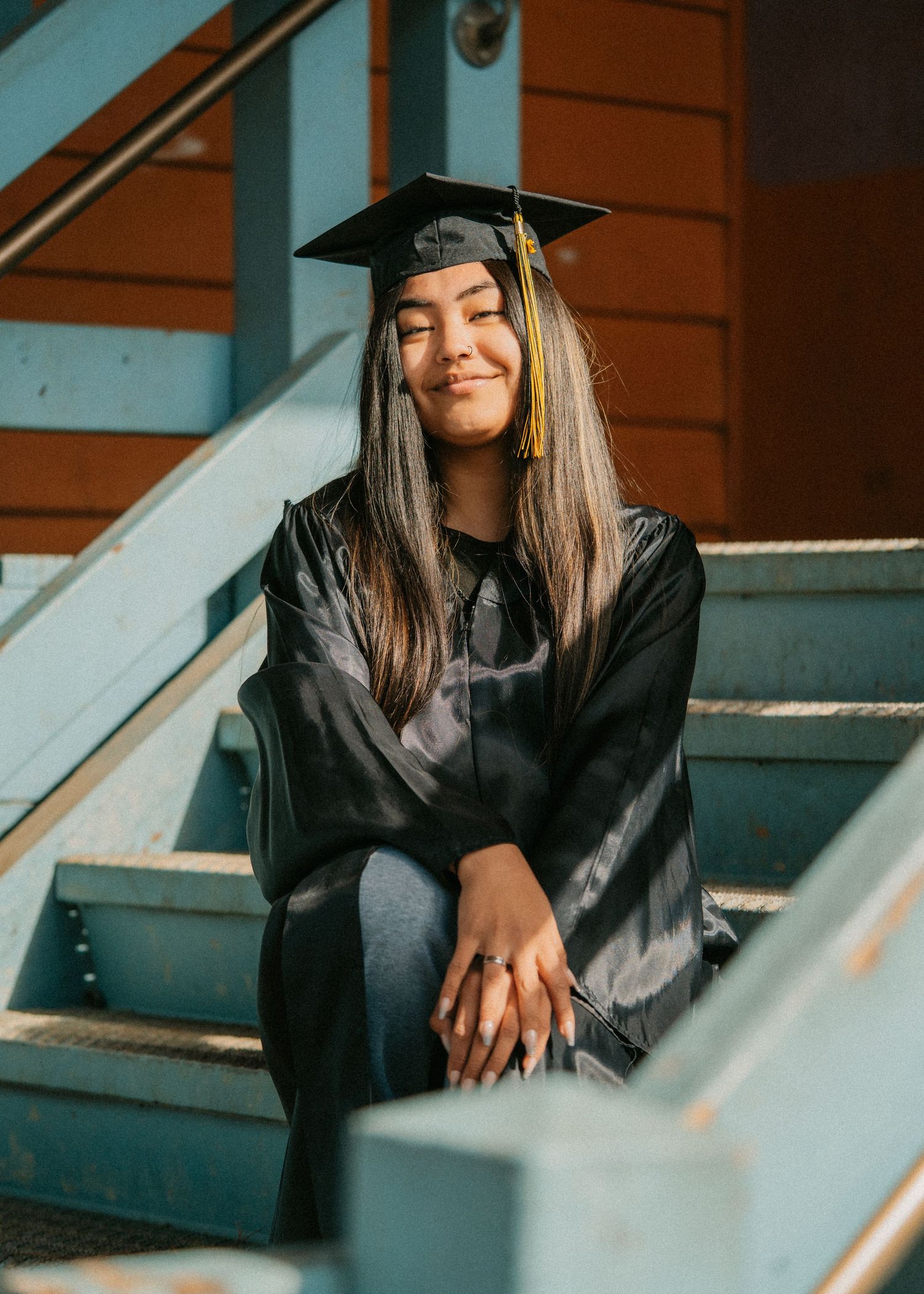 Female graduate in black cap and gown sitting peacefully on blue outdoor stairs in sunlight. Anchorage-Senior-Photographer