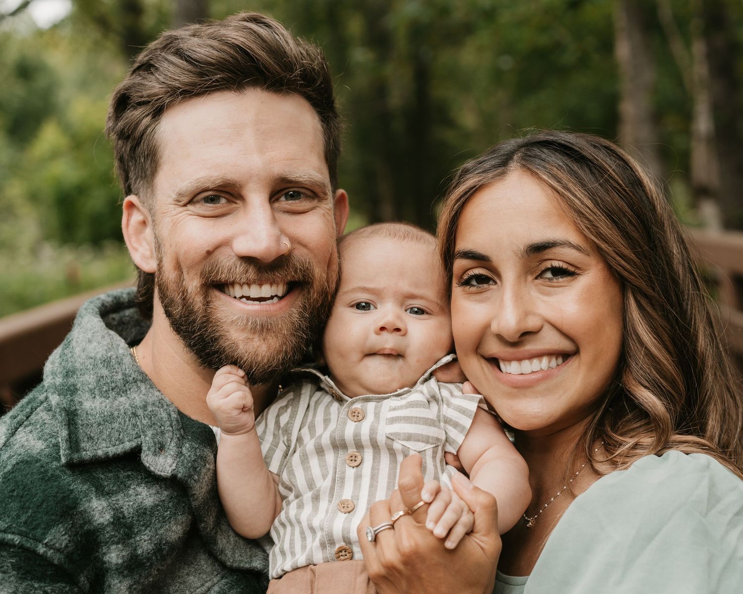 Smiling couple holding their newborn baby outdoors surrounded by lush green trees in a warm portrait. anchorage-family-photographer