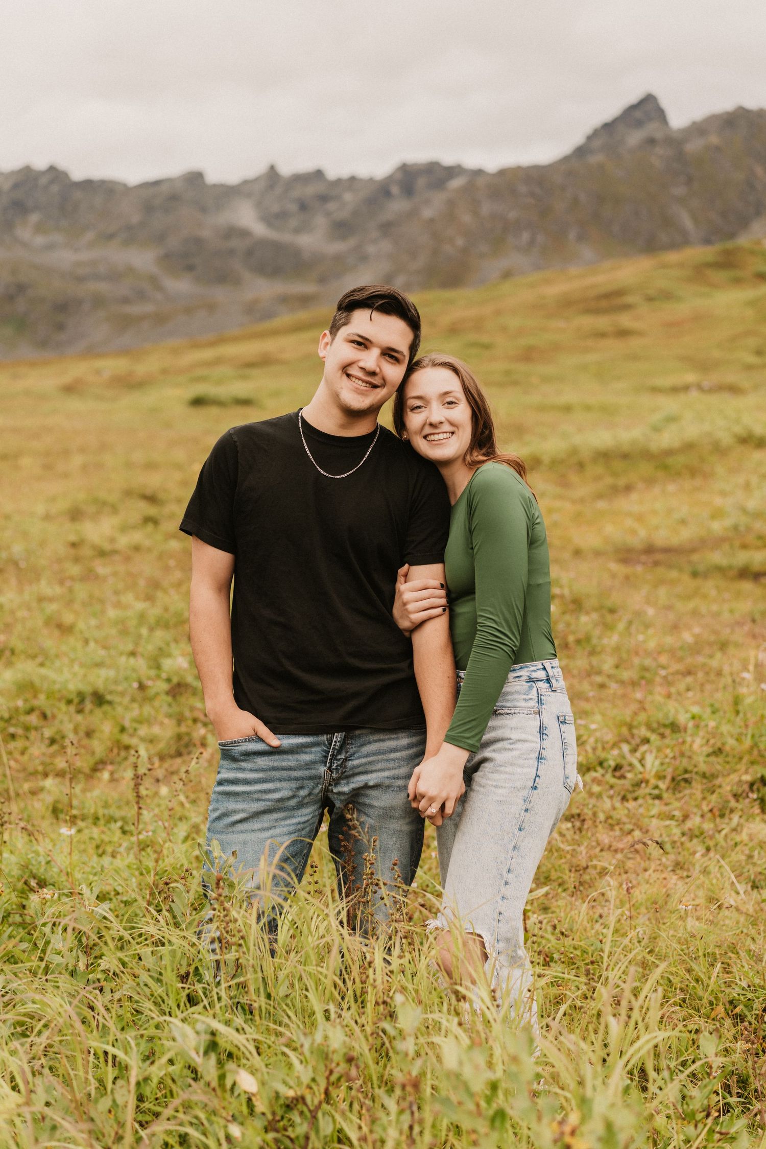 A smiling couple embraces in a grassy alpine meadow with Rocky Mountains in the background. Anchorage-Couple-Photographer