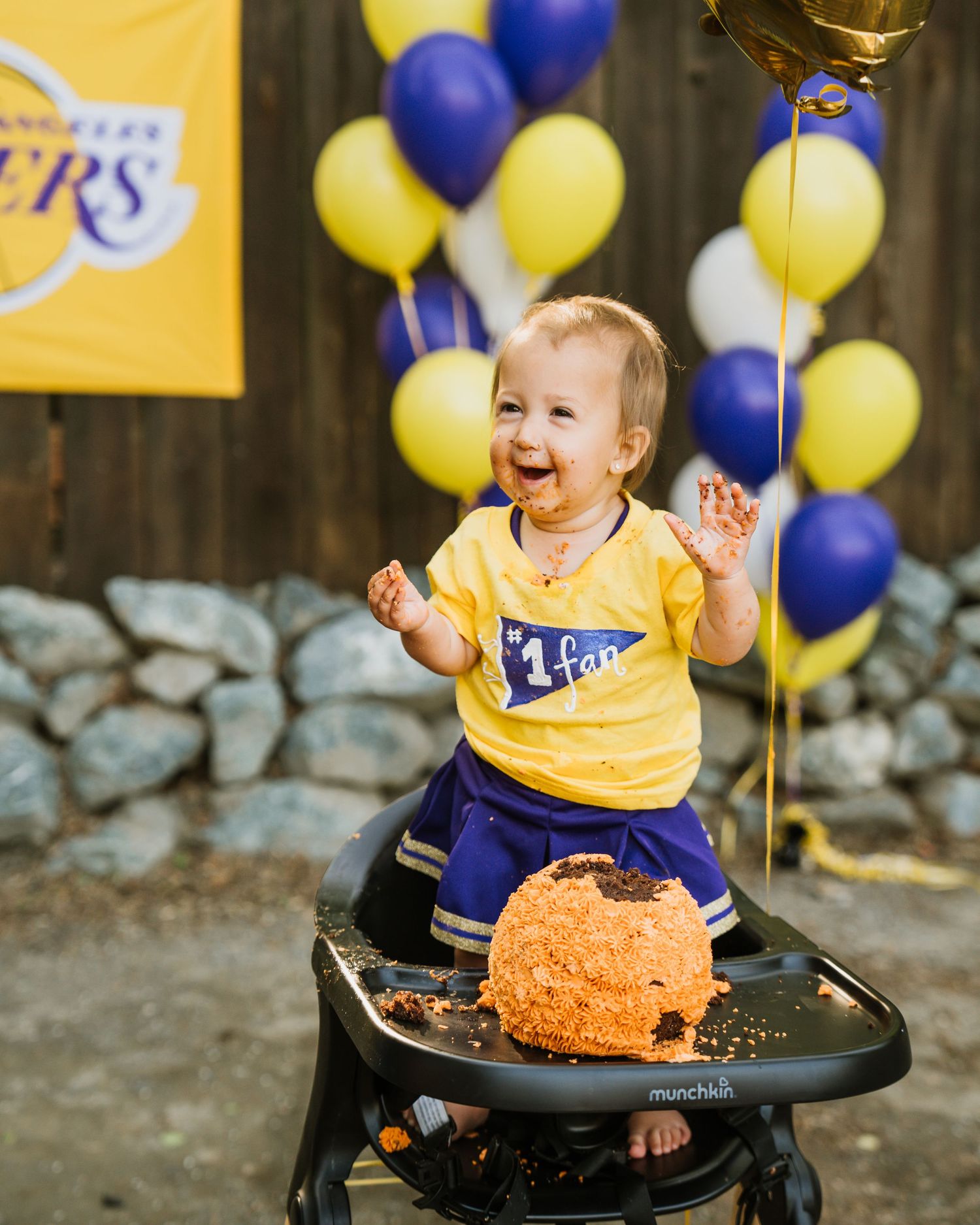 Smiling baby in Lakers jersey celebrates first birthday cake smash with colourful balloons behind. anchorage-event-photographer