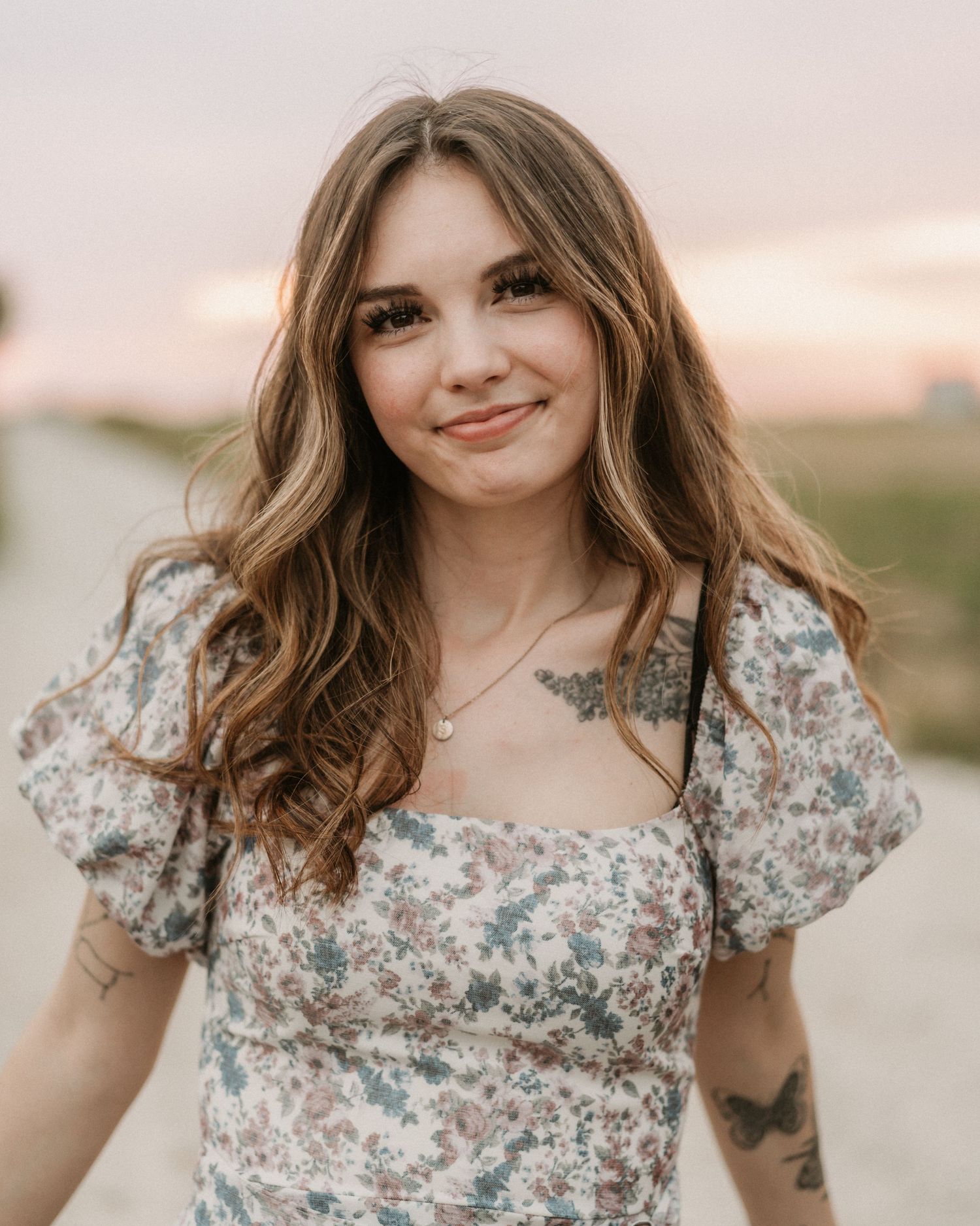 Young woman with long brown hair and tattoos smiling outdoors in a floral dress at sunset.  Anchorage-Senior-Photographer