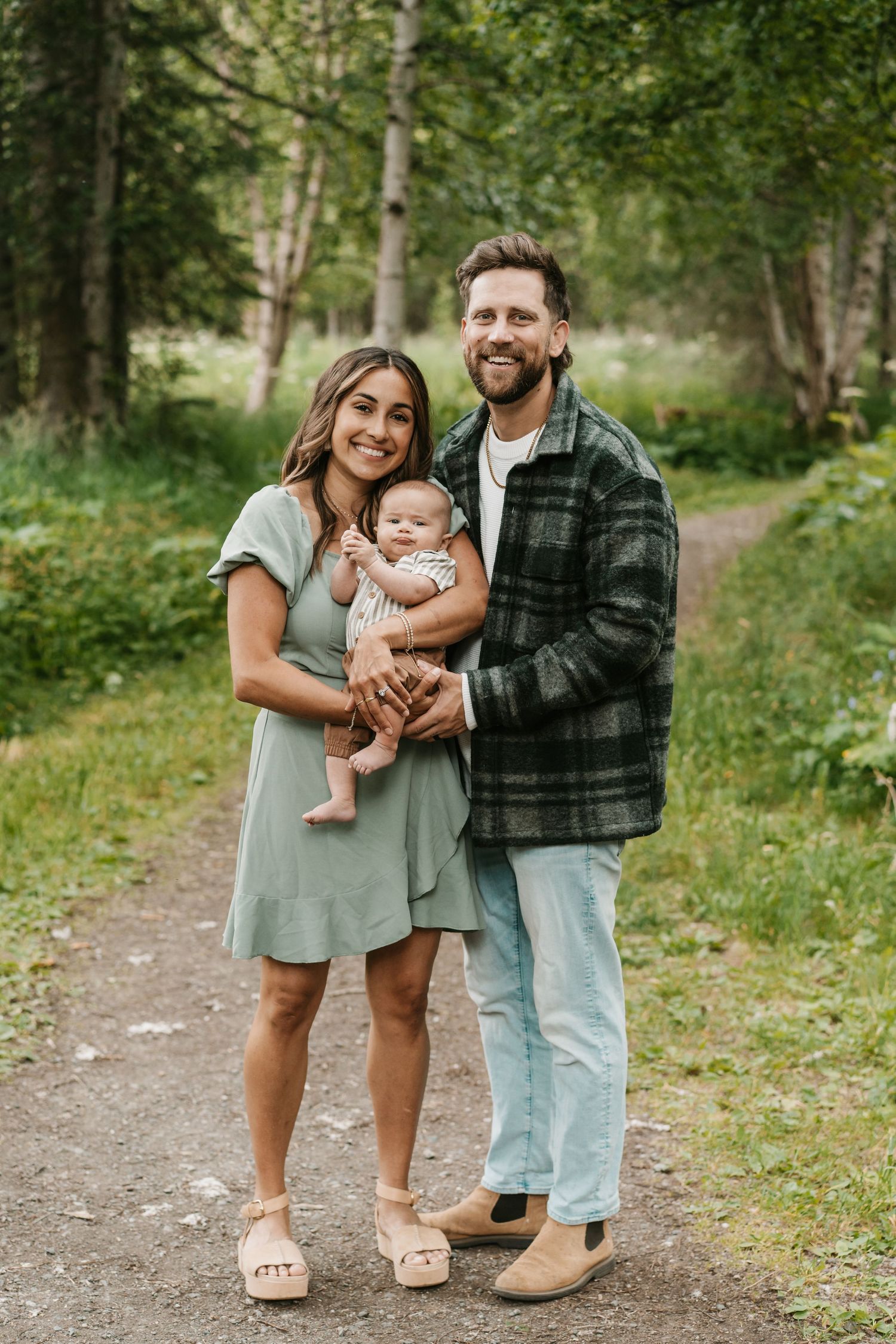 Smiling family of three stands on a forest path, mother in green dress holding baby, father in plaid. anchorage-family-photographer