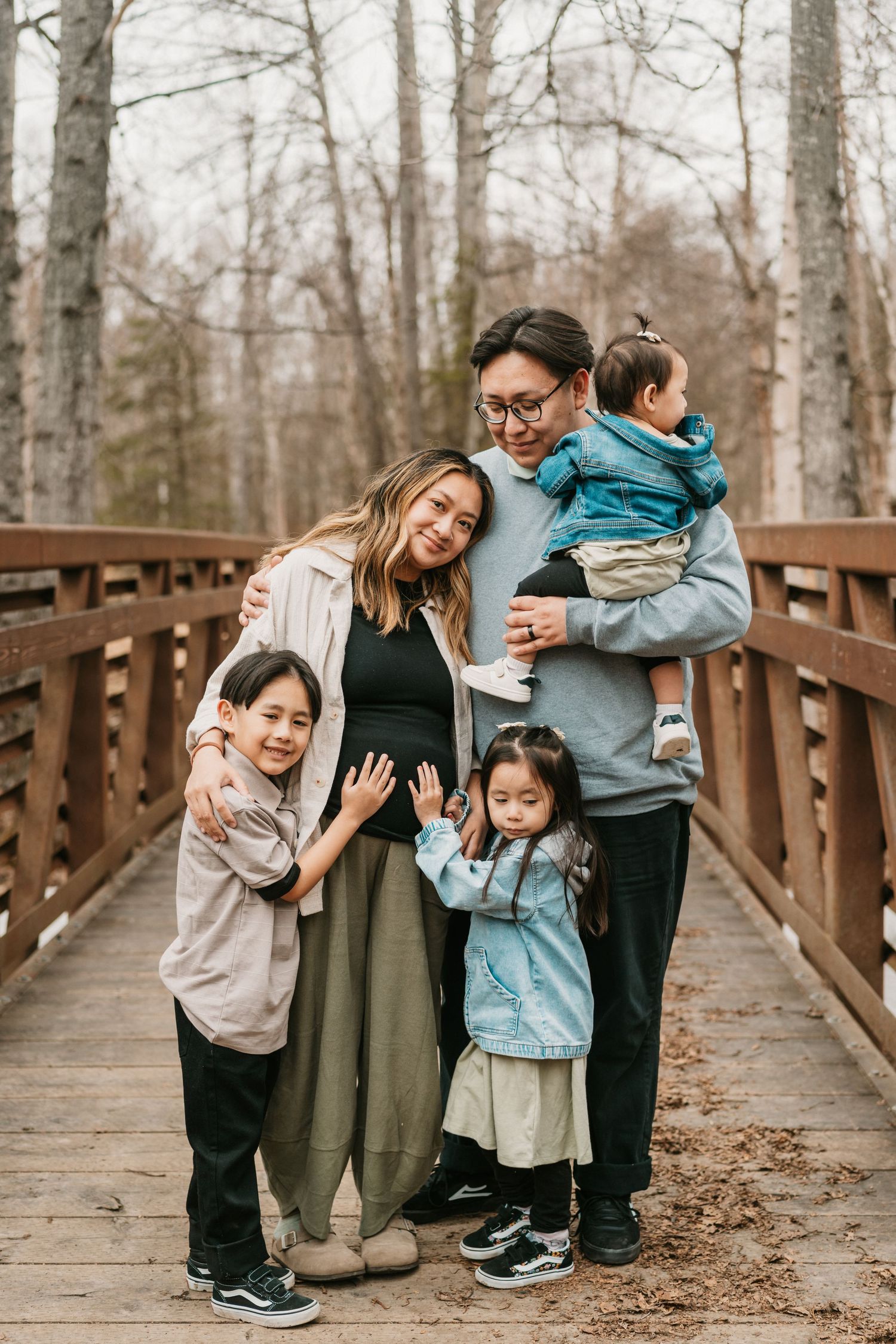 Asian family of five poses on wooden bridge in forest, pregnant mother surrounded by husband and three young children.