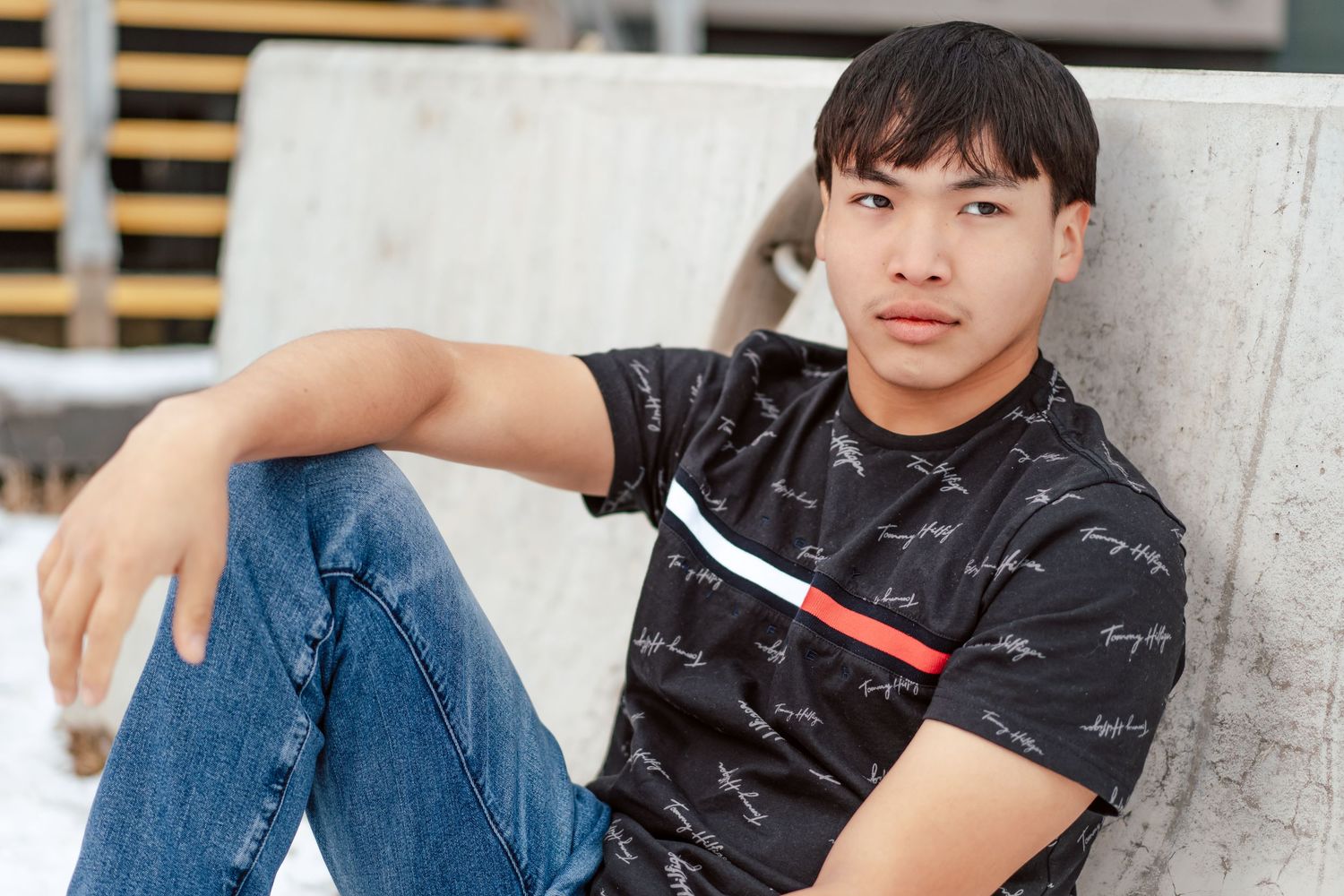 Young Asian man in black graphic tee and jeans sitting casually against a concrete wall outdoors.