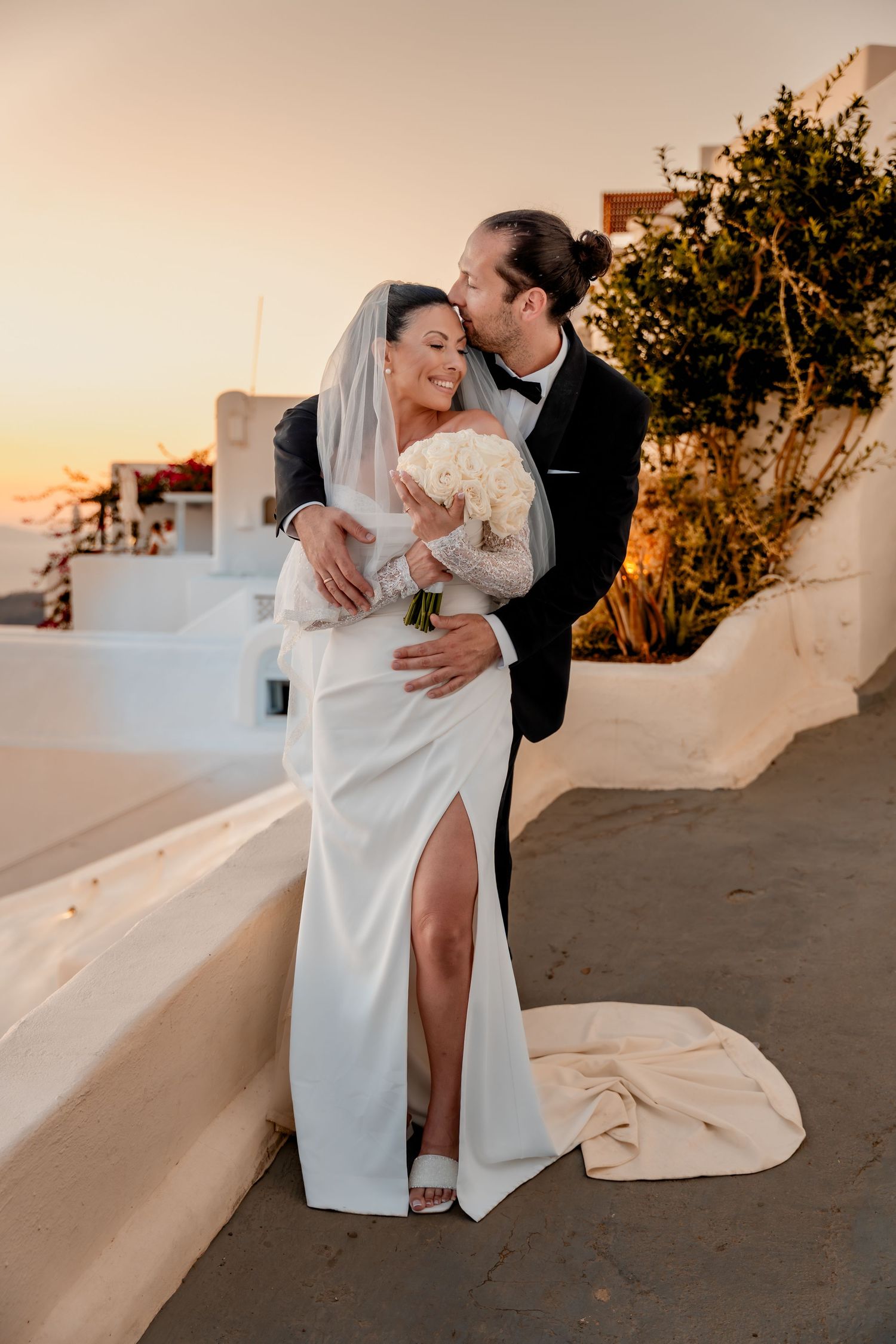 Newlyweds kissing on a terrace in Santorini, sunset and flowers in the background 🌅