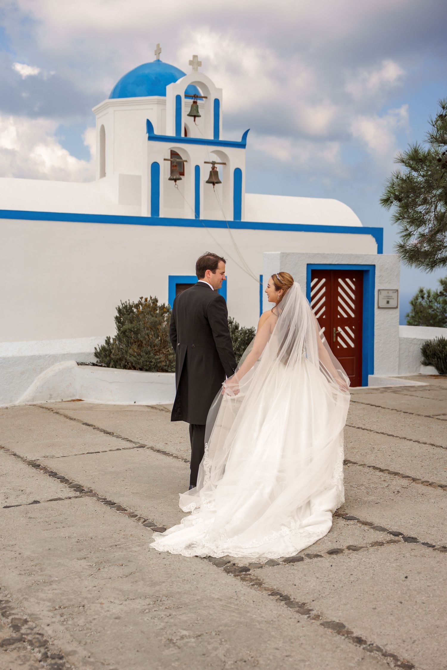 Bride in wedding dress, groom in suit, captured on Santorini cliffs walking hand in hand after their wedding  ceremony