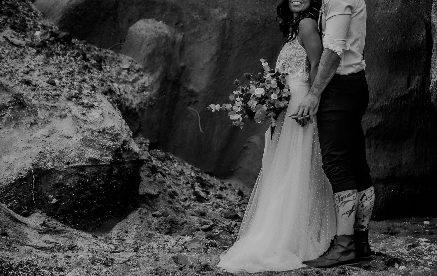 Intimate elopement photoshoot of a couple at Santorini's wild lava rock formations, with flowers and natural light. Bride in a wedding dress