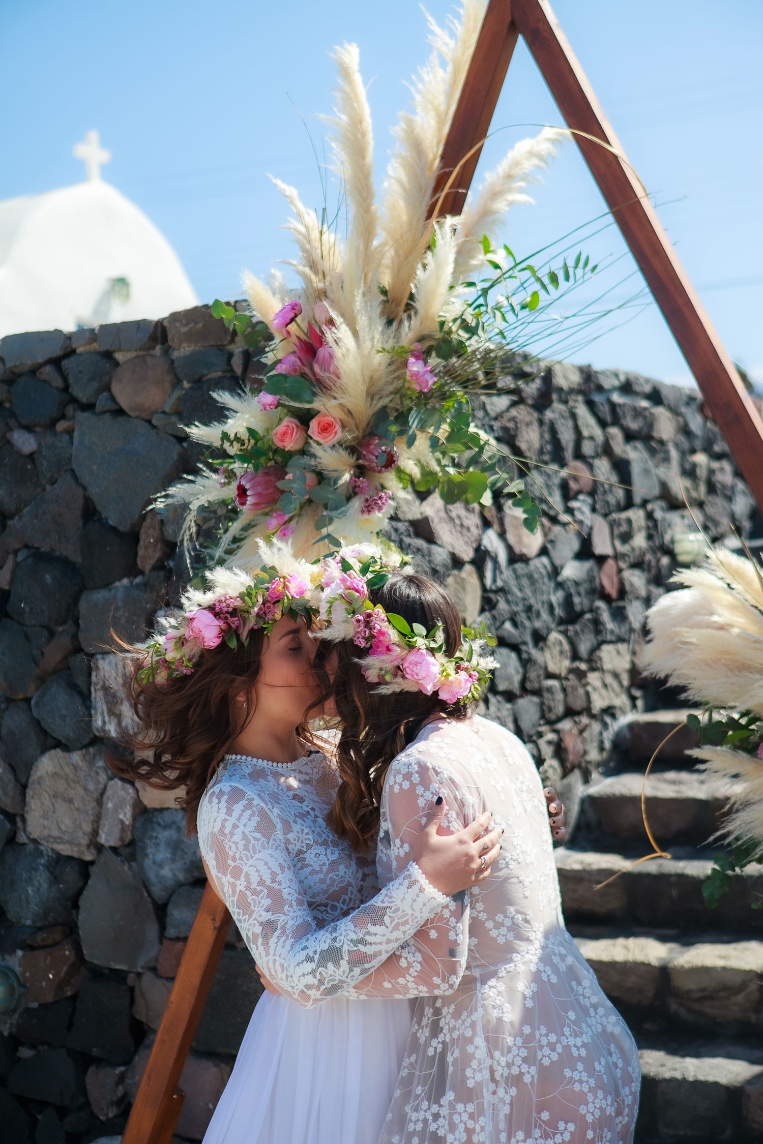 Two women in flower crowns kissing by floral decor on a Santorini cliff, with black stone fence and traditional church in background.