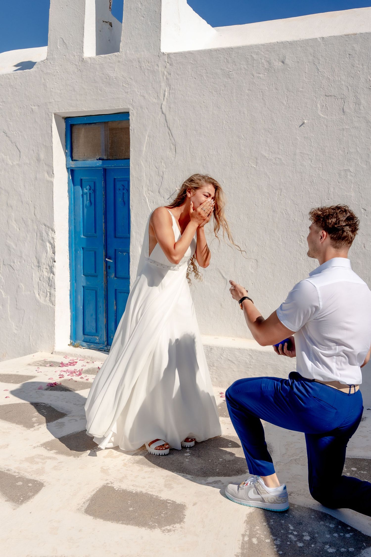 Groom on one knee, bride smiling during a Santorini proposal, love in every glance