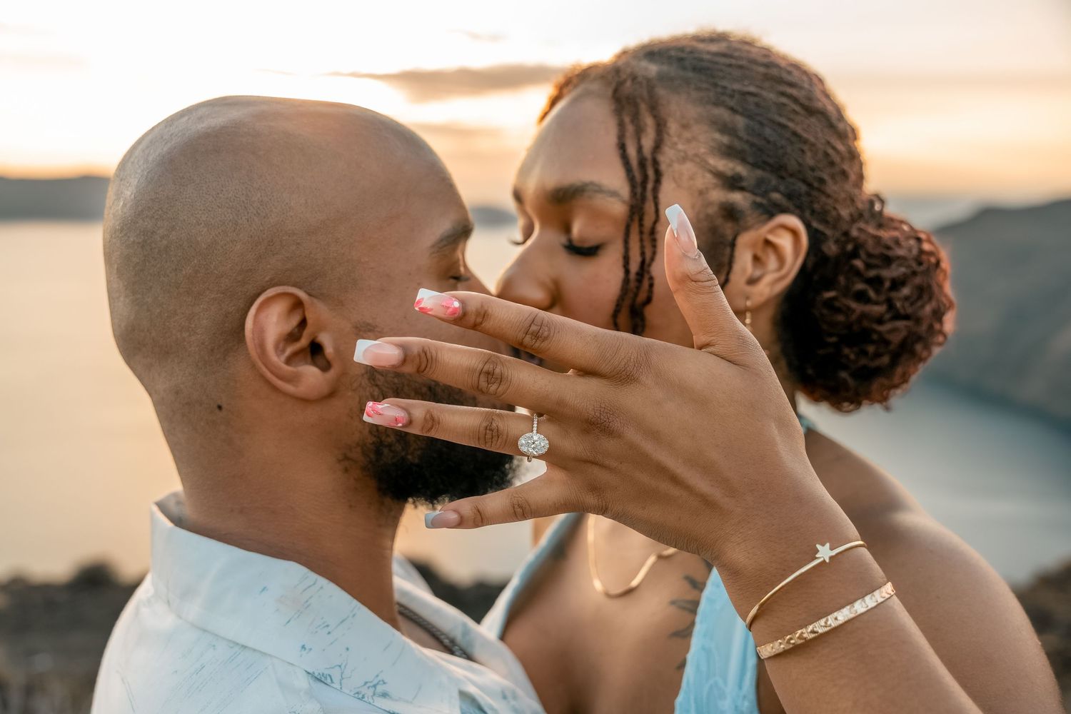 Couple kissing, bride saying yes and showing her wedding ring, embracing an intimate proposal on Santorini cliffs during the golden hour.