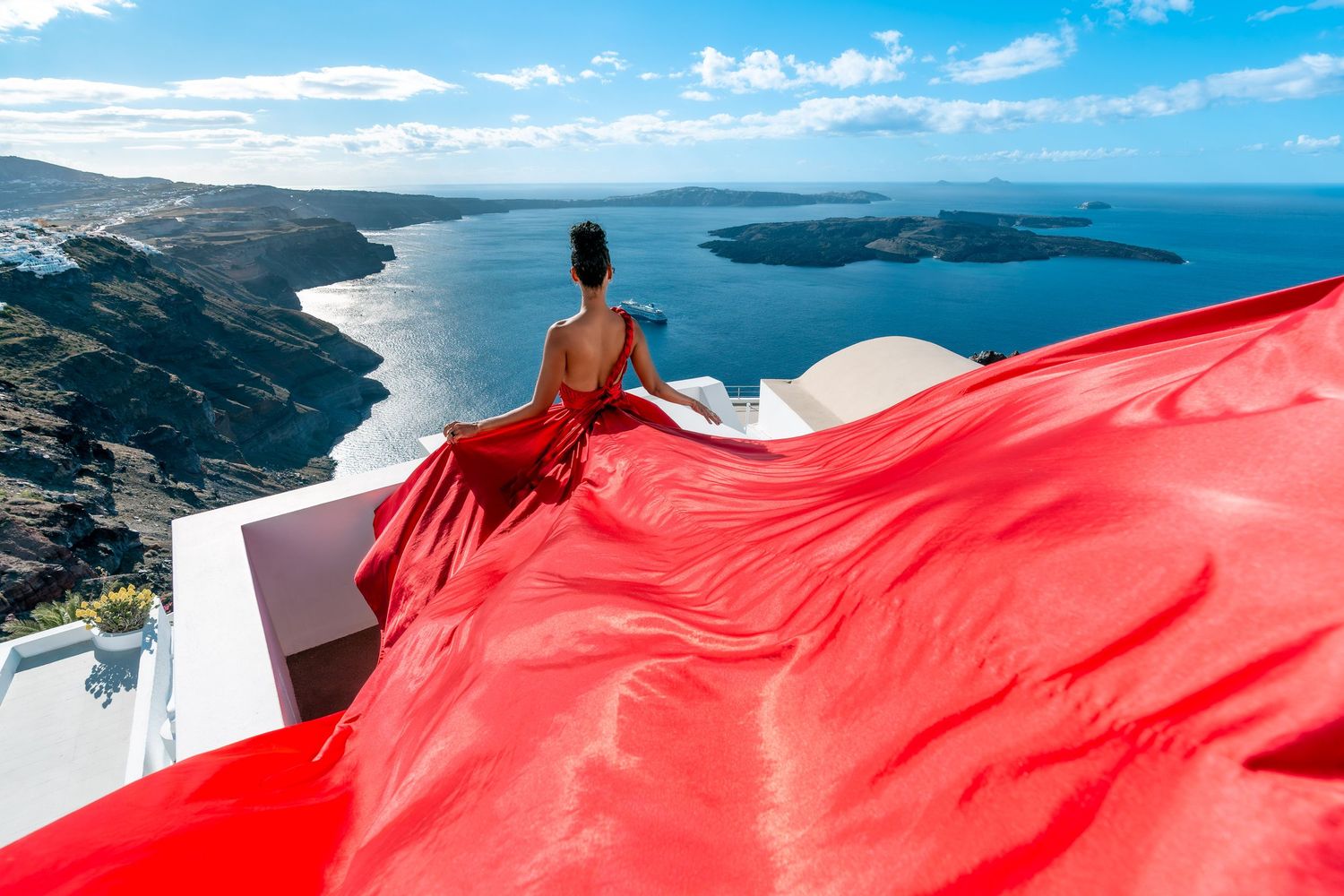 Fashionable and sensual lifestyle photoshoot of a woman in beautiful red flying dress on a Santorini terrace, natural light, and happiness.