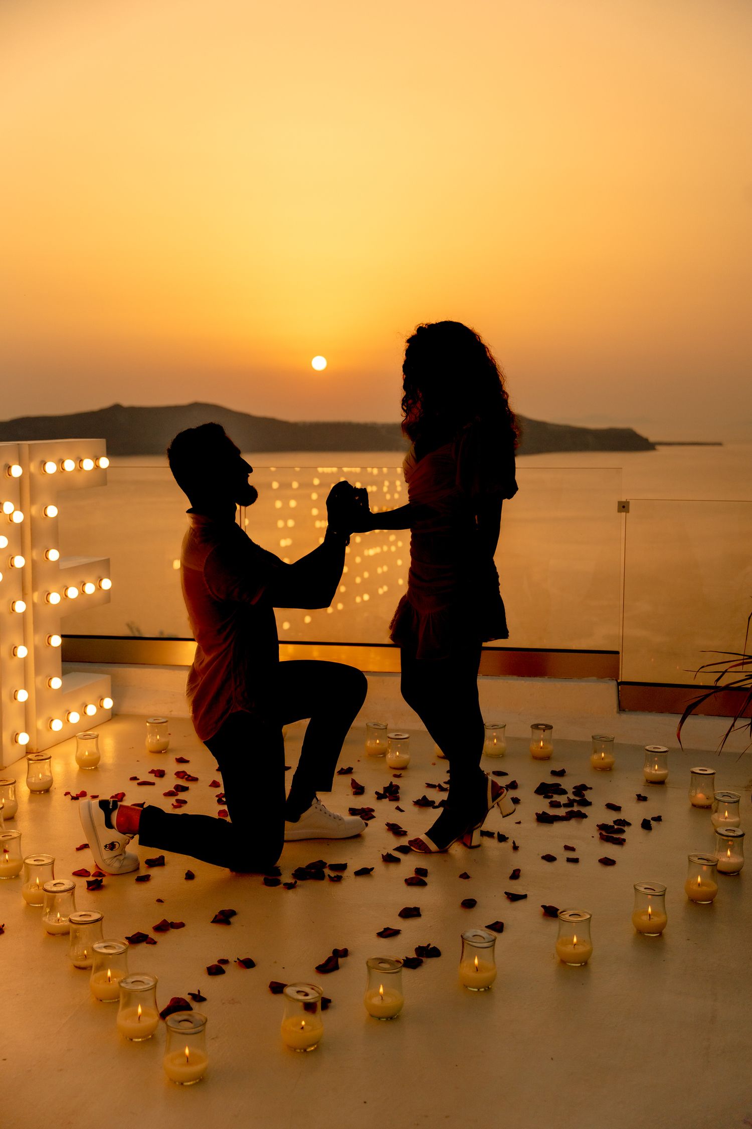 Groom on his knee proposing to bride on a Santorini terrace at sunset, capturing love and excitement 🌅