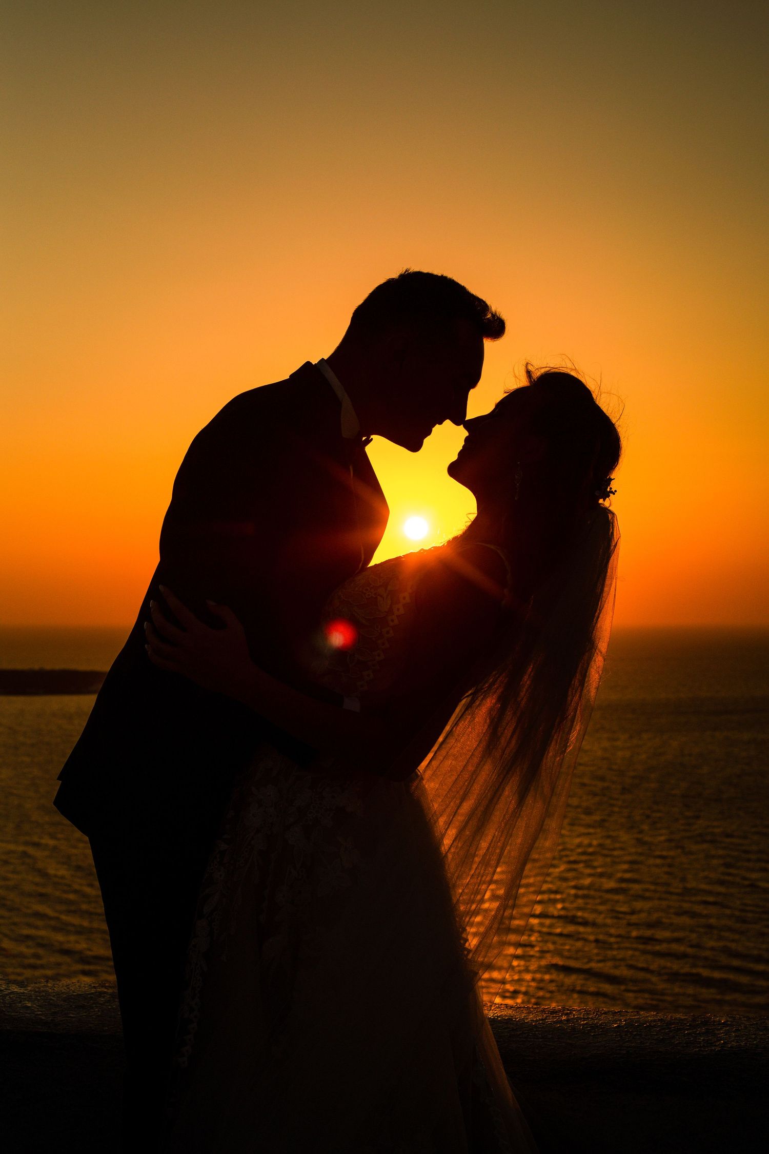 Newlyweds kissing on a terrace in Santorini, sunset and flowers in the background 🌅