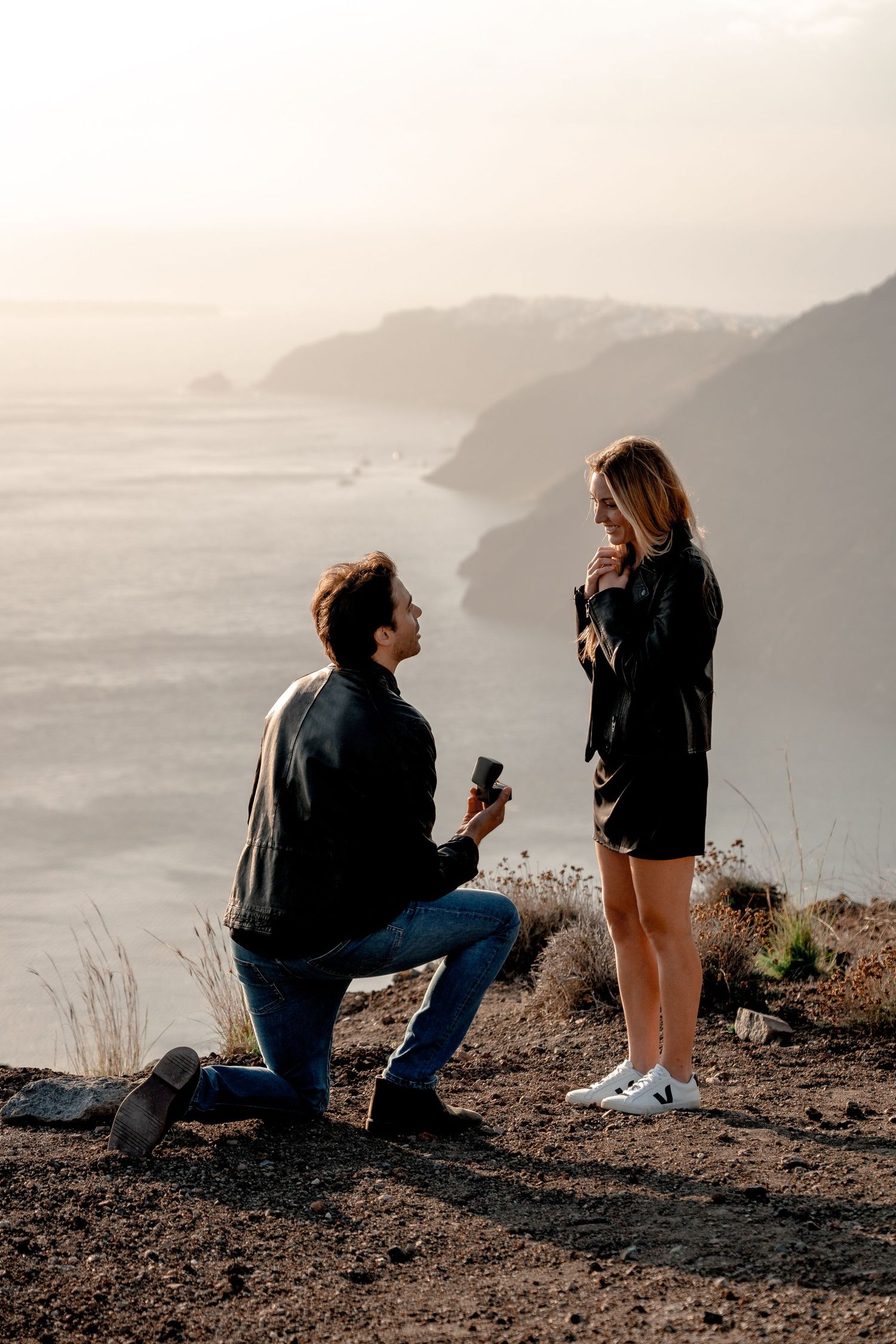 Groom proposing to bride on a Santorini cliffside at sunset, capturing love and excitement 🌅