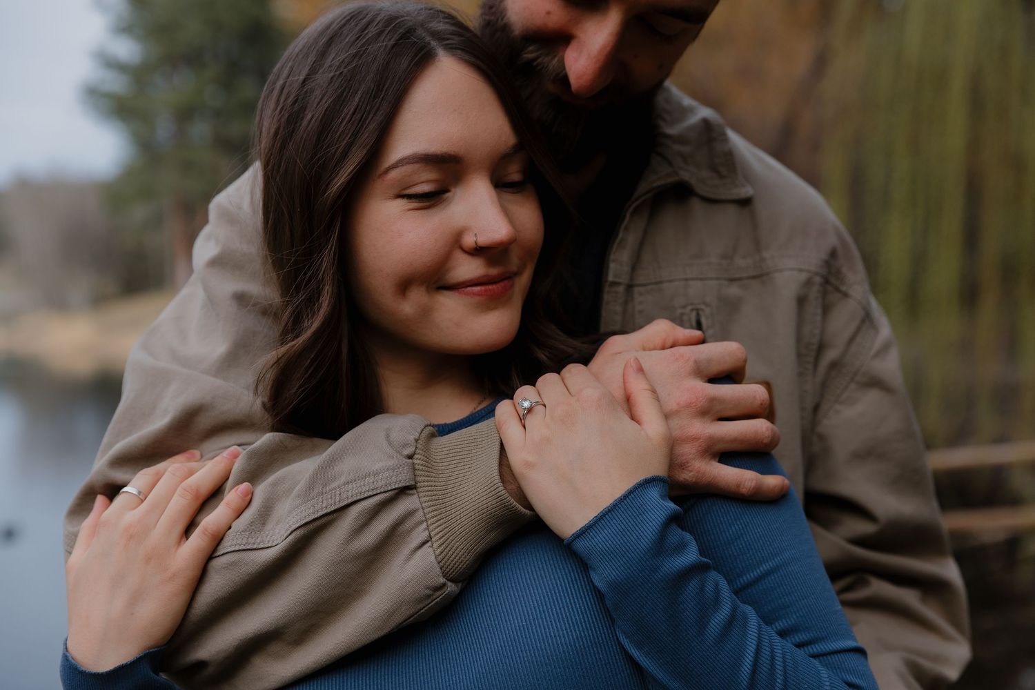 A woman smiling contentedly as her partner wraps his arms around her near a serene pond.