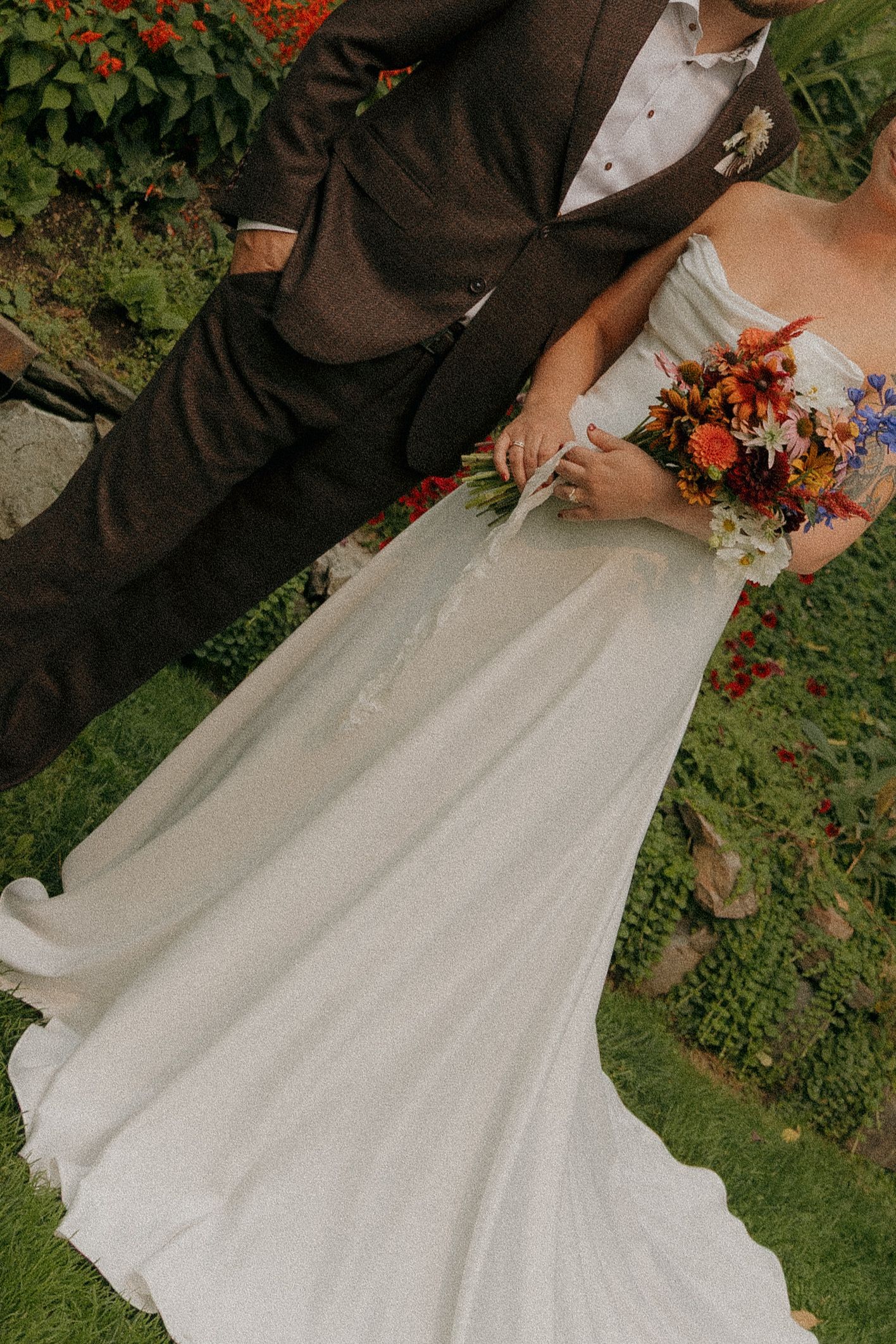 Bride in white gown holds colorful bouquet beside groom in brown suit in lush garden setting.