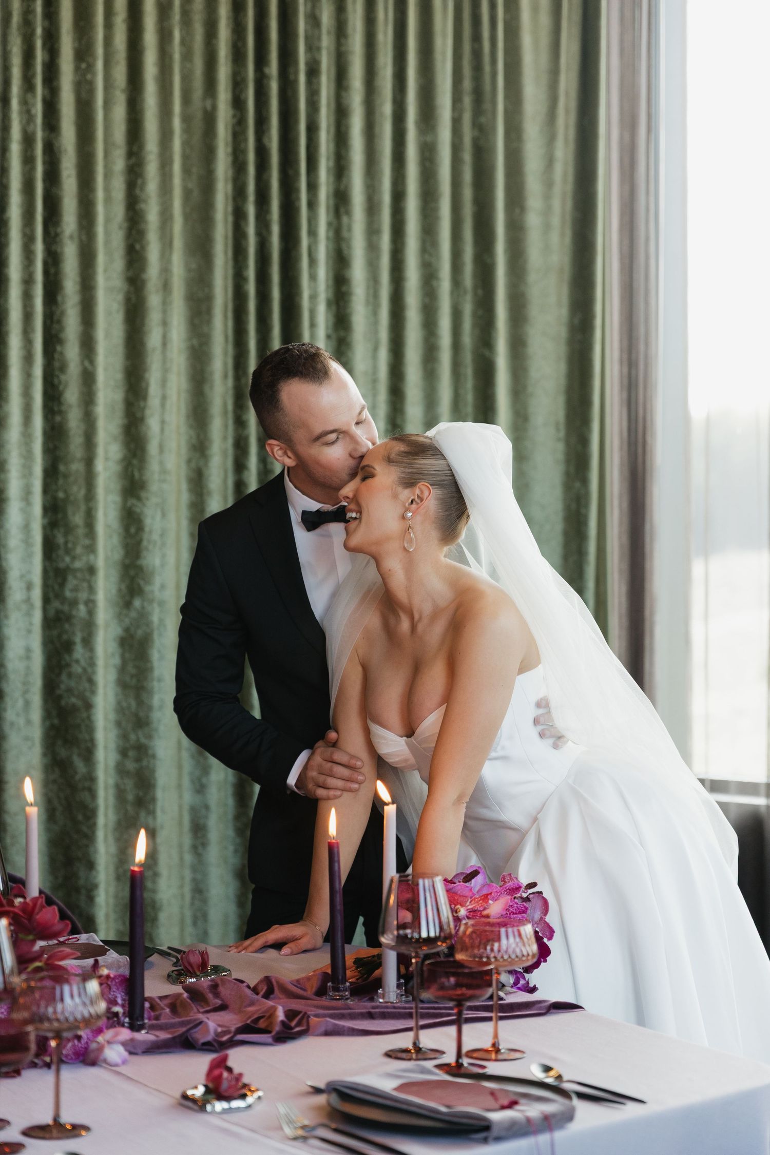 Groom in black tuxedo kisses bride in white strapless gown at candlelit wedding reception table with purple florals.