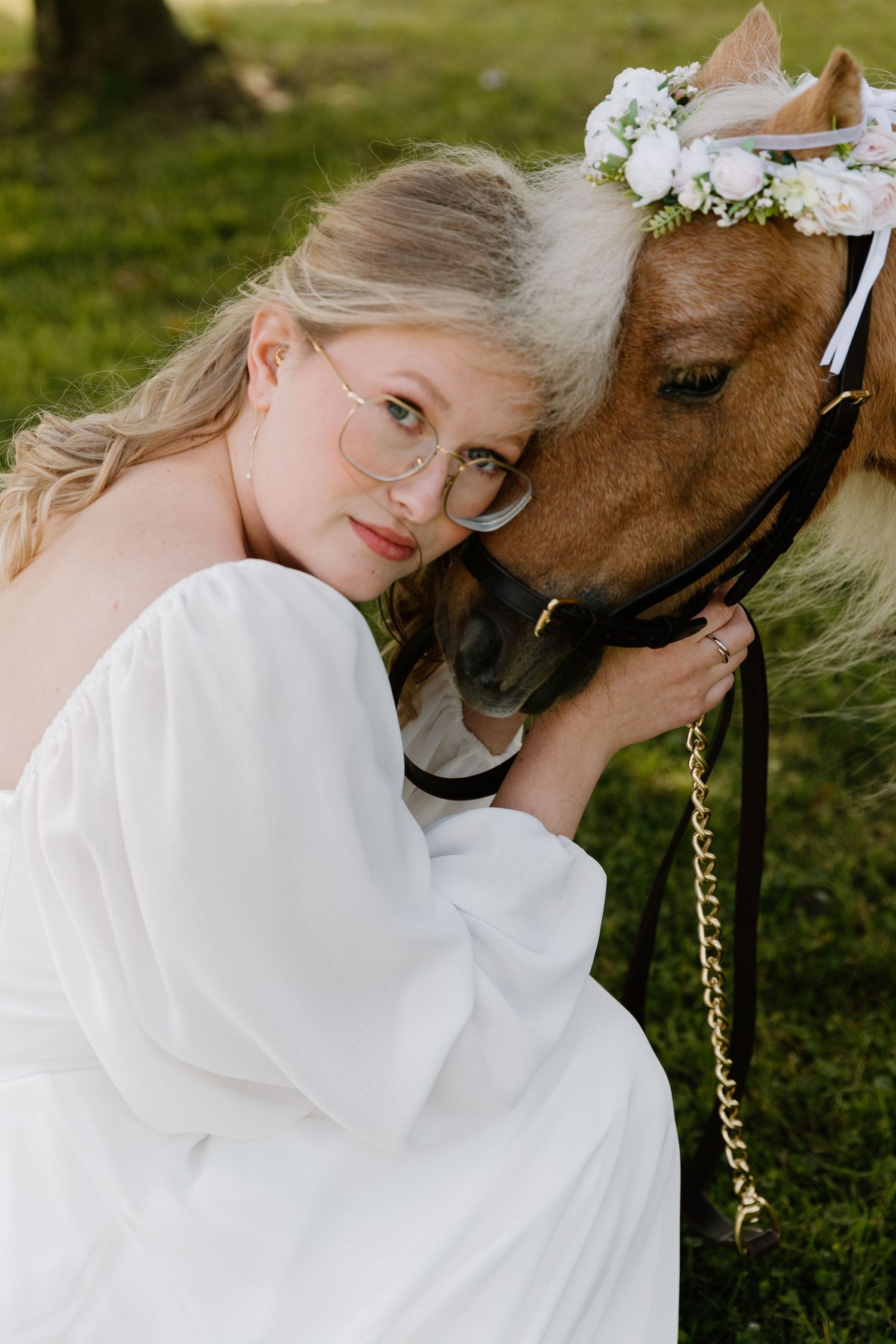 Bride in white gown and glasses tenderly embraces a brown pony wearing a floral crown outdoors.