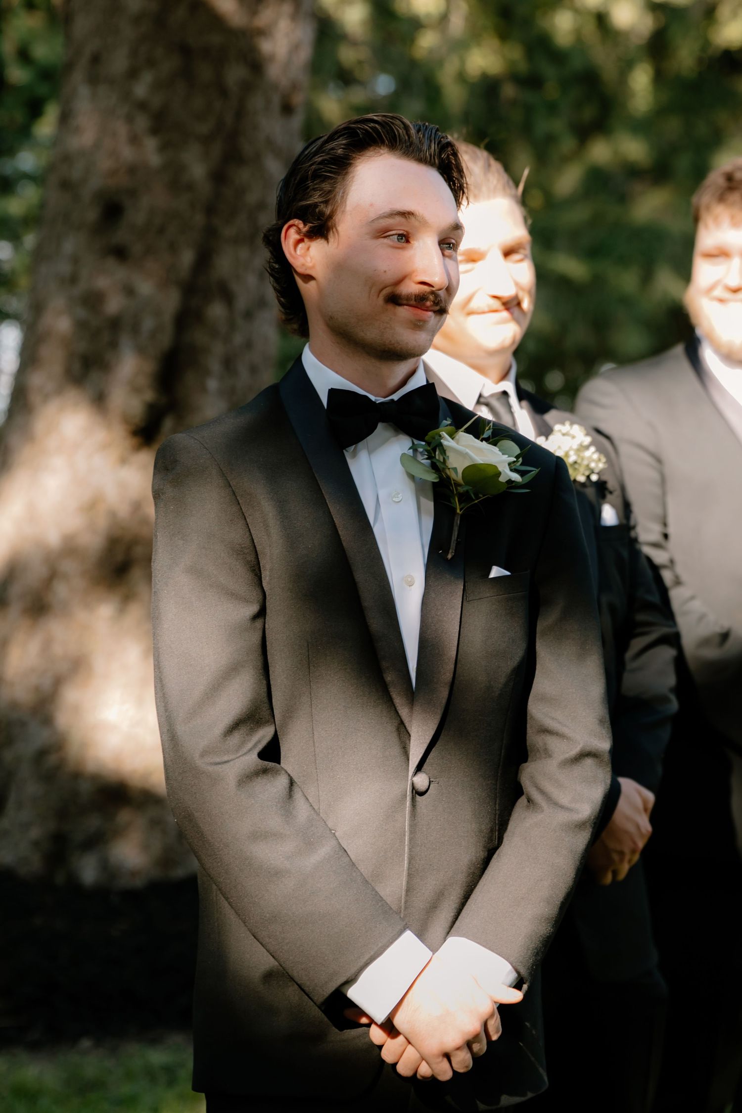Groom in grey tuxedo with bow tie and boutonniere waiting at outdoor wedding ceremony altar.