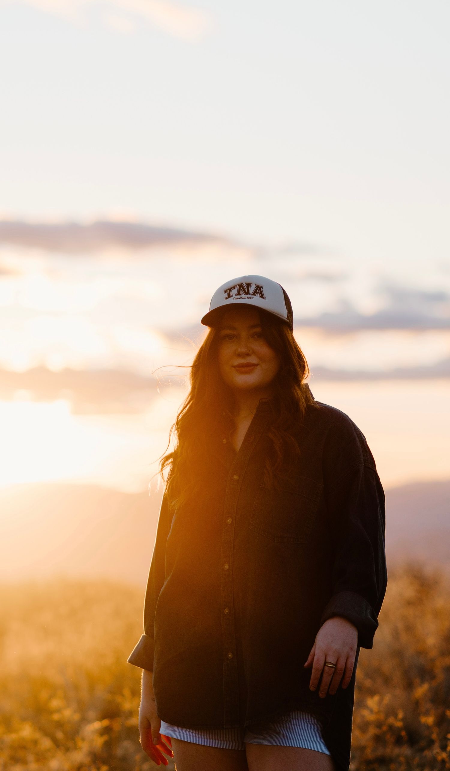Woman in TNA trucker hat posing at sunset with glowing sky behind her.