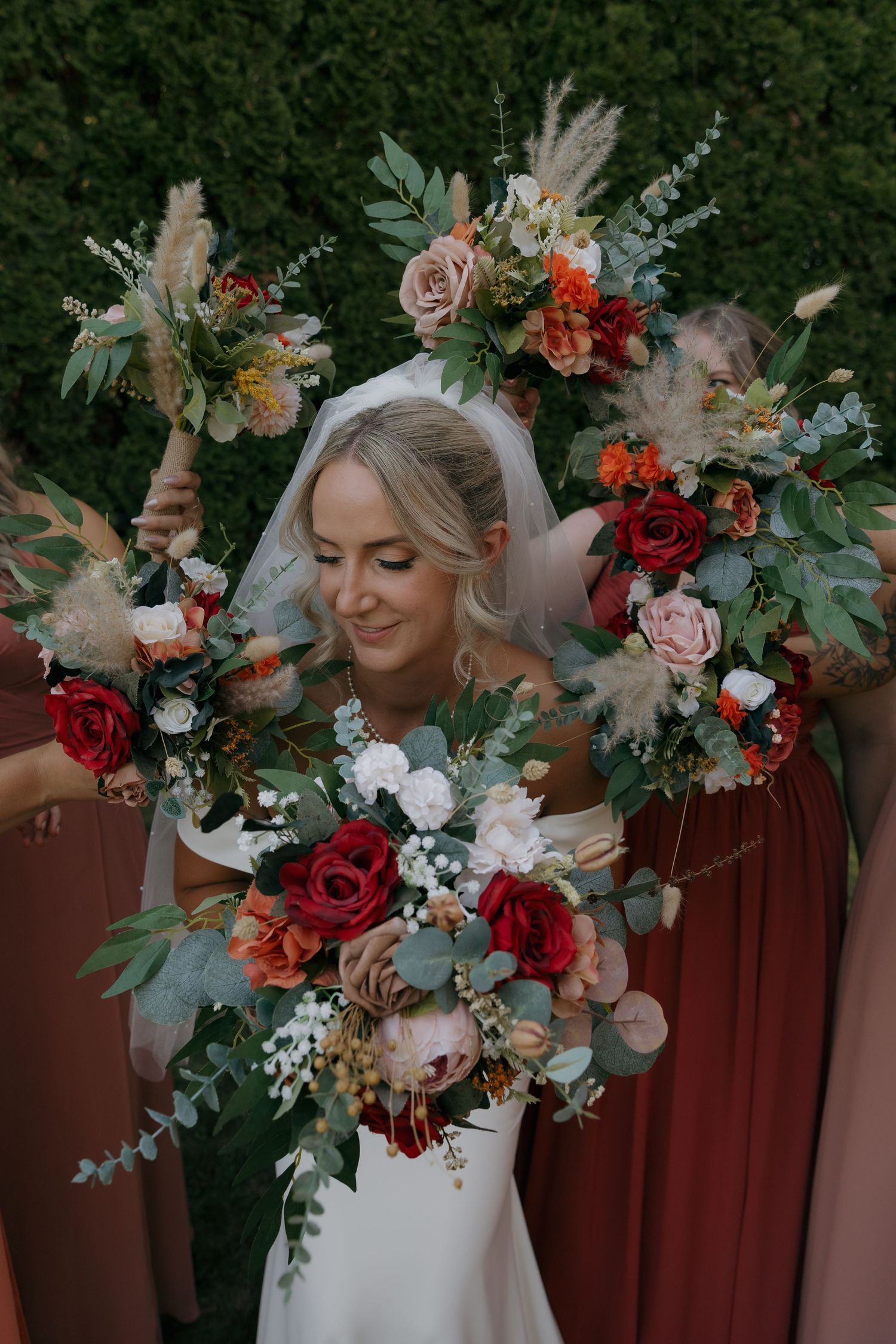 Bride surrounded by bridesmaids holding colorful bouquets of red roses and eucalyptus outdoors.