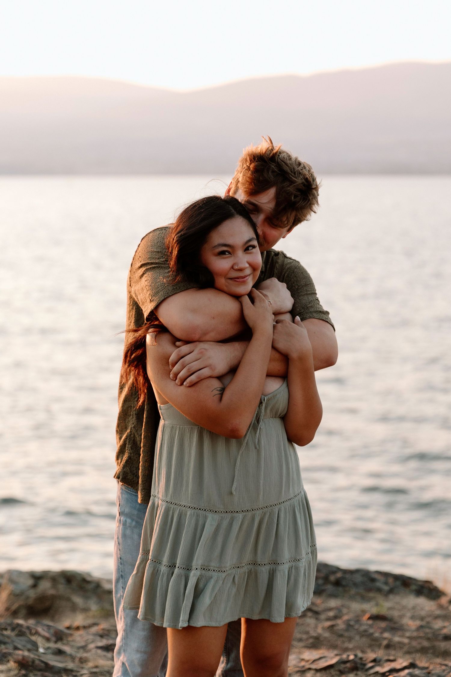 Man hugging woman from behind on rocky shoreline at sunset with calm lake behind them.