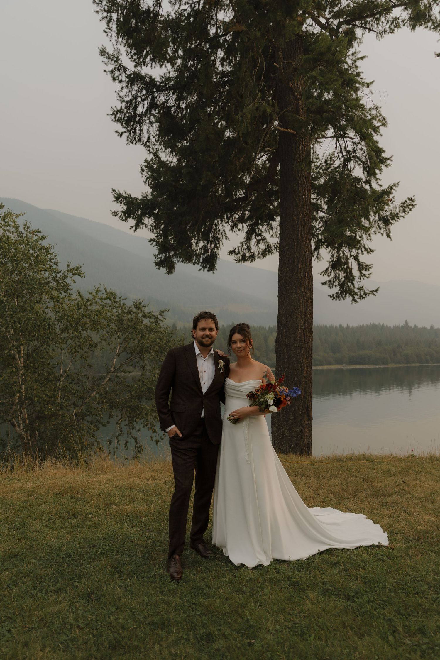 Bride and groom posing under a tall pine tree beside a misty mountain lake.
