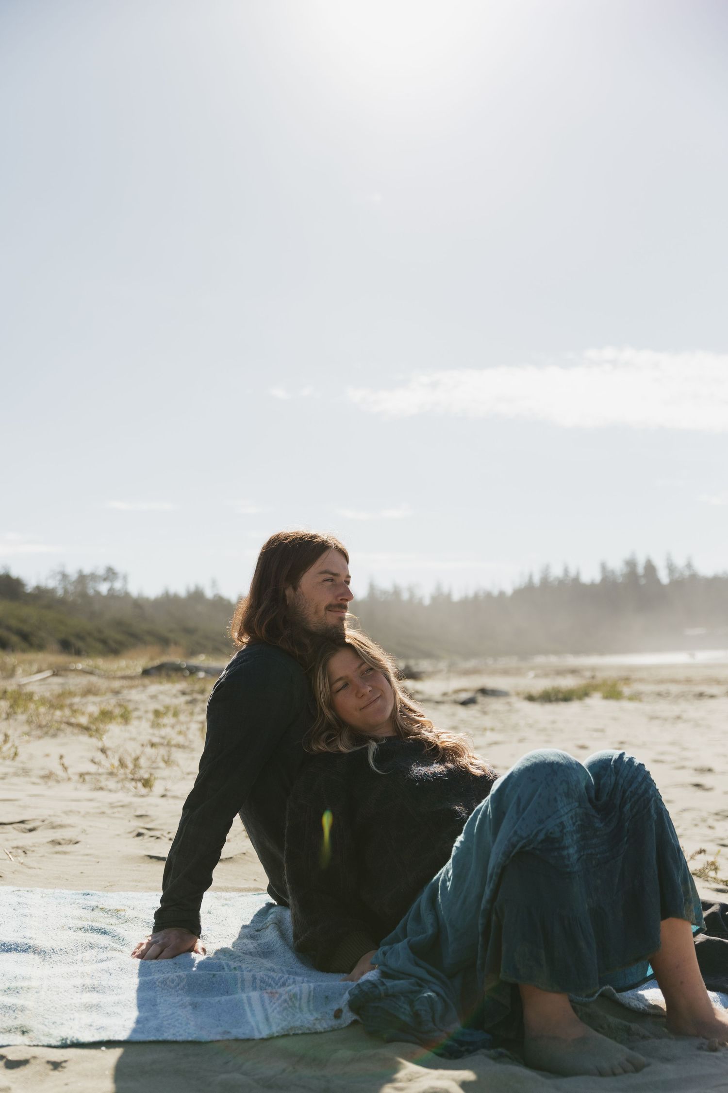 Couple sitting together on a sunny beach, woman resting her head on mans shoulder.