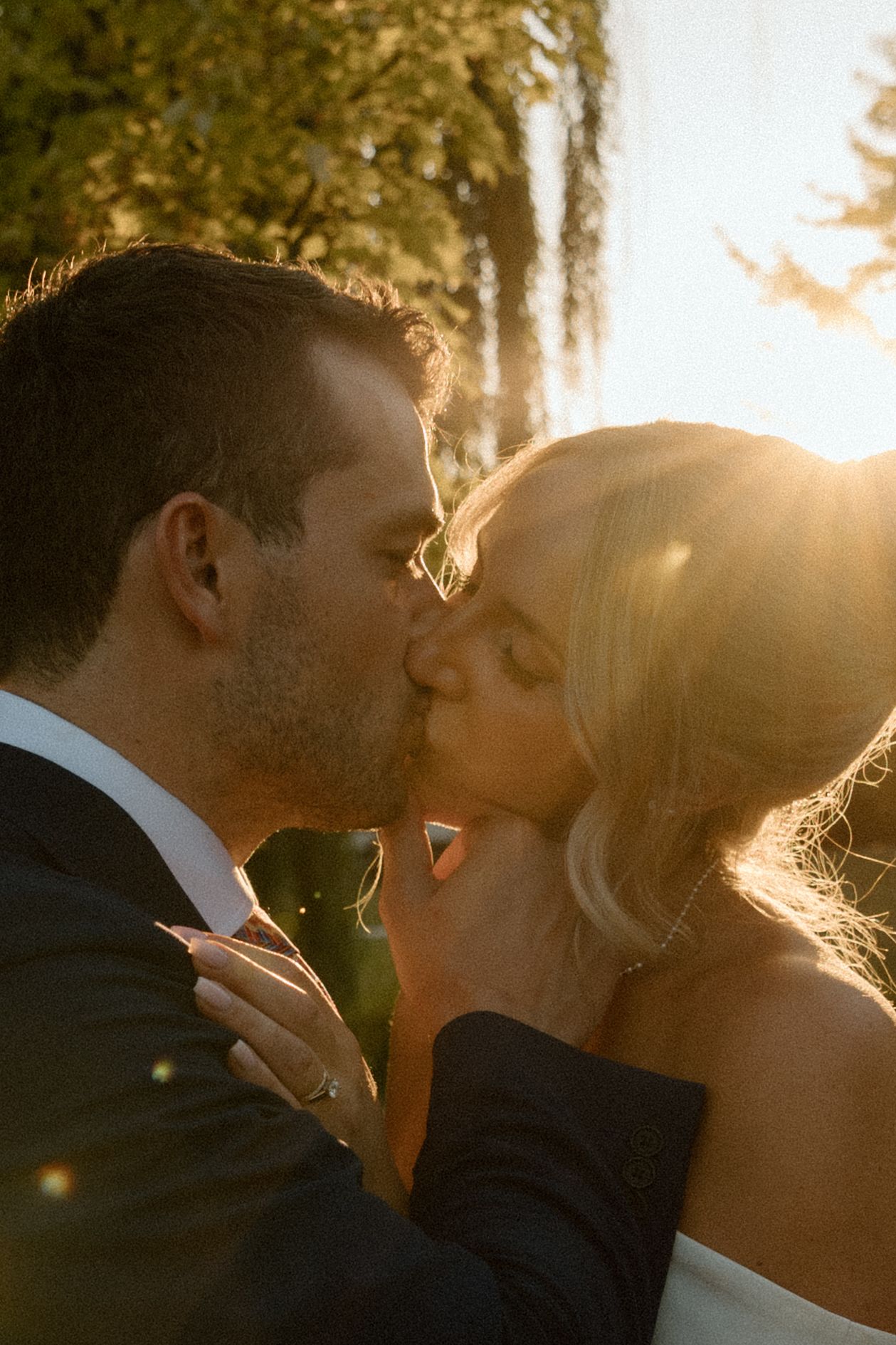 Bride and groom share a romantic kiss bathed in golden sunset light outdoors.