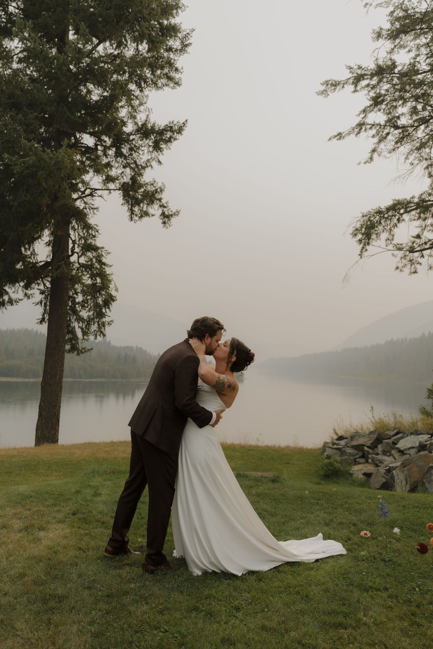 Bride and groom share first kiss by a misty lake surrounded by tall pine trees.