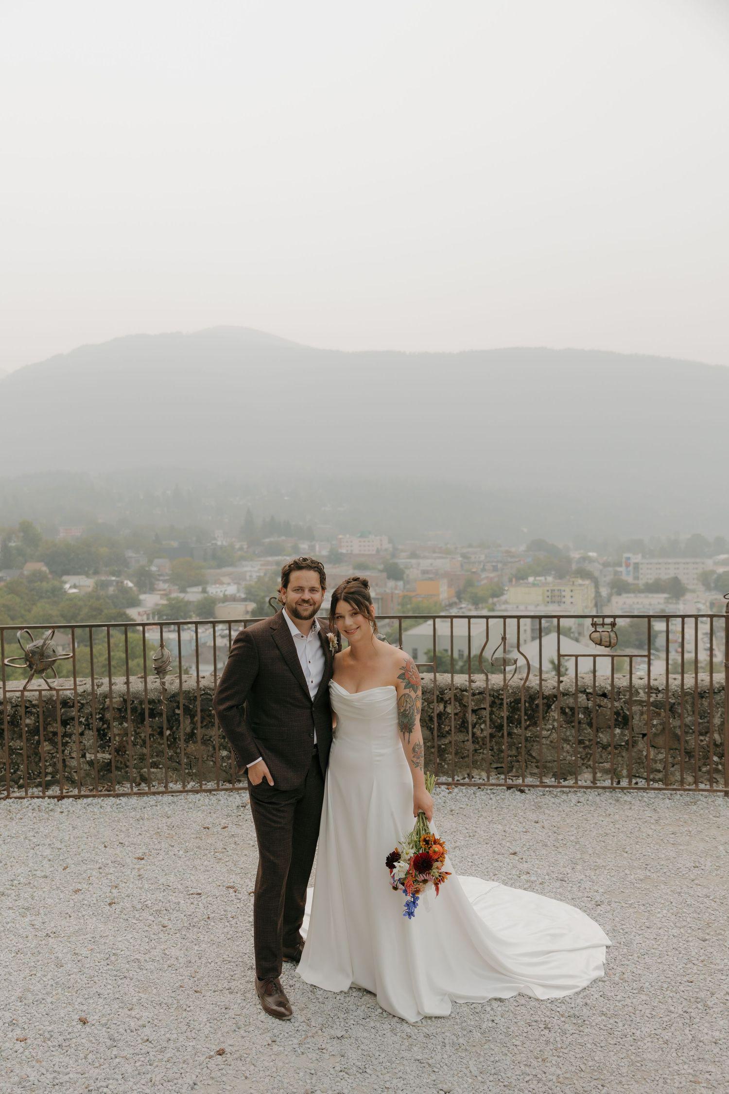 Bride and groom pose on stone terrace with misty mountain town backdrop on wedding day.