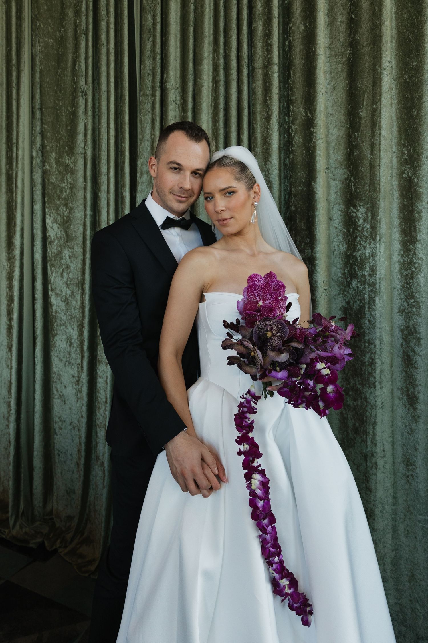 Bride and groom pose elegantly before green velvet drapes, bride holding dramatic cascading purple orchid bouquet.