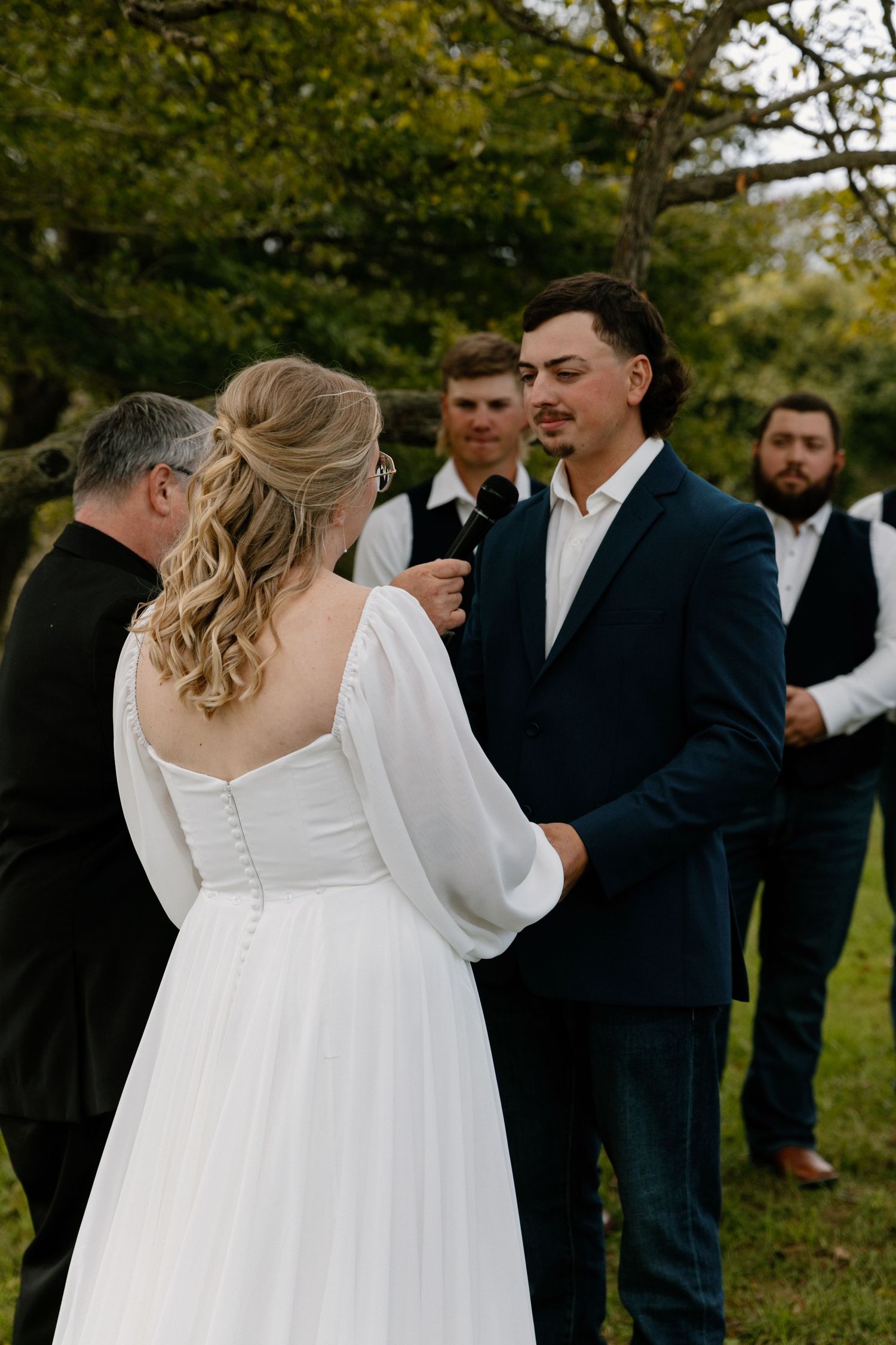 Groom in navy suit holds bride's hands during outdoor wedding ceremony with groomsmen behind.