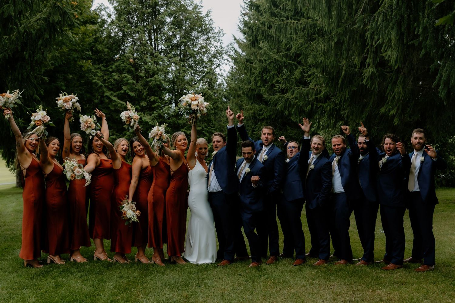 Wedding party posing outdoors with bridesmaids in rust orange dresses and groomsmen in navy suits.