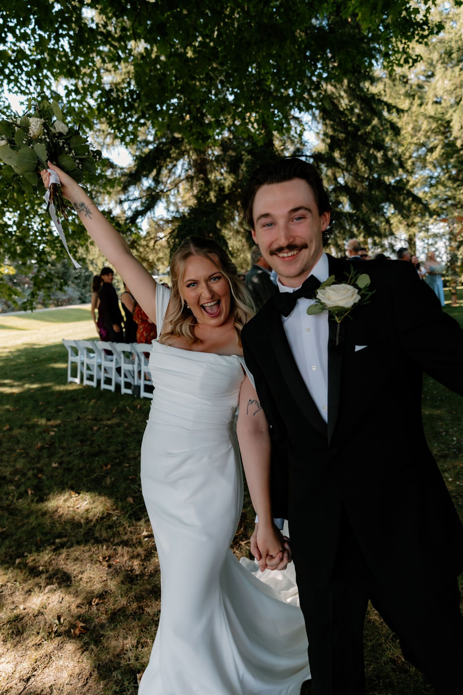 Jubilant bride and groom celebrating just-married moment under tree after outdoor wedding ceremony.