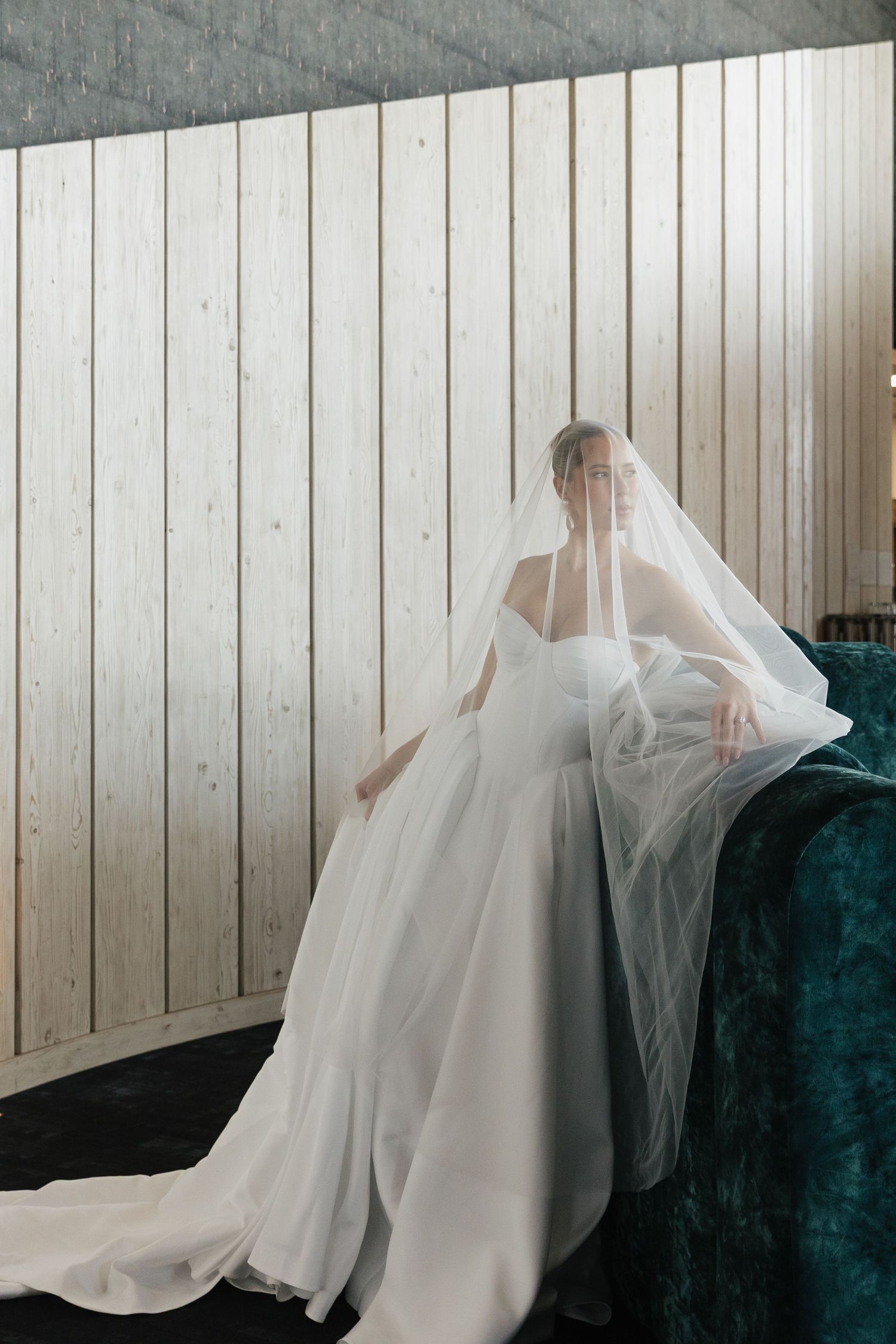 Bride in flowing white strapless gown and long veil poses gracefully against a teal velvet sofa indoors.