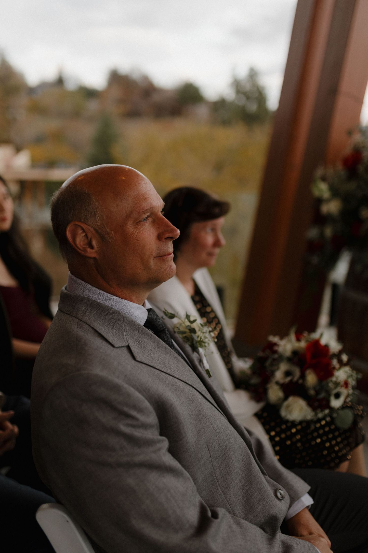 Smiling male wedding guest in gray suit with boutonniere seated during outdoor wedding ceremony.