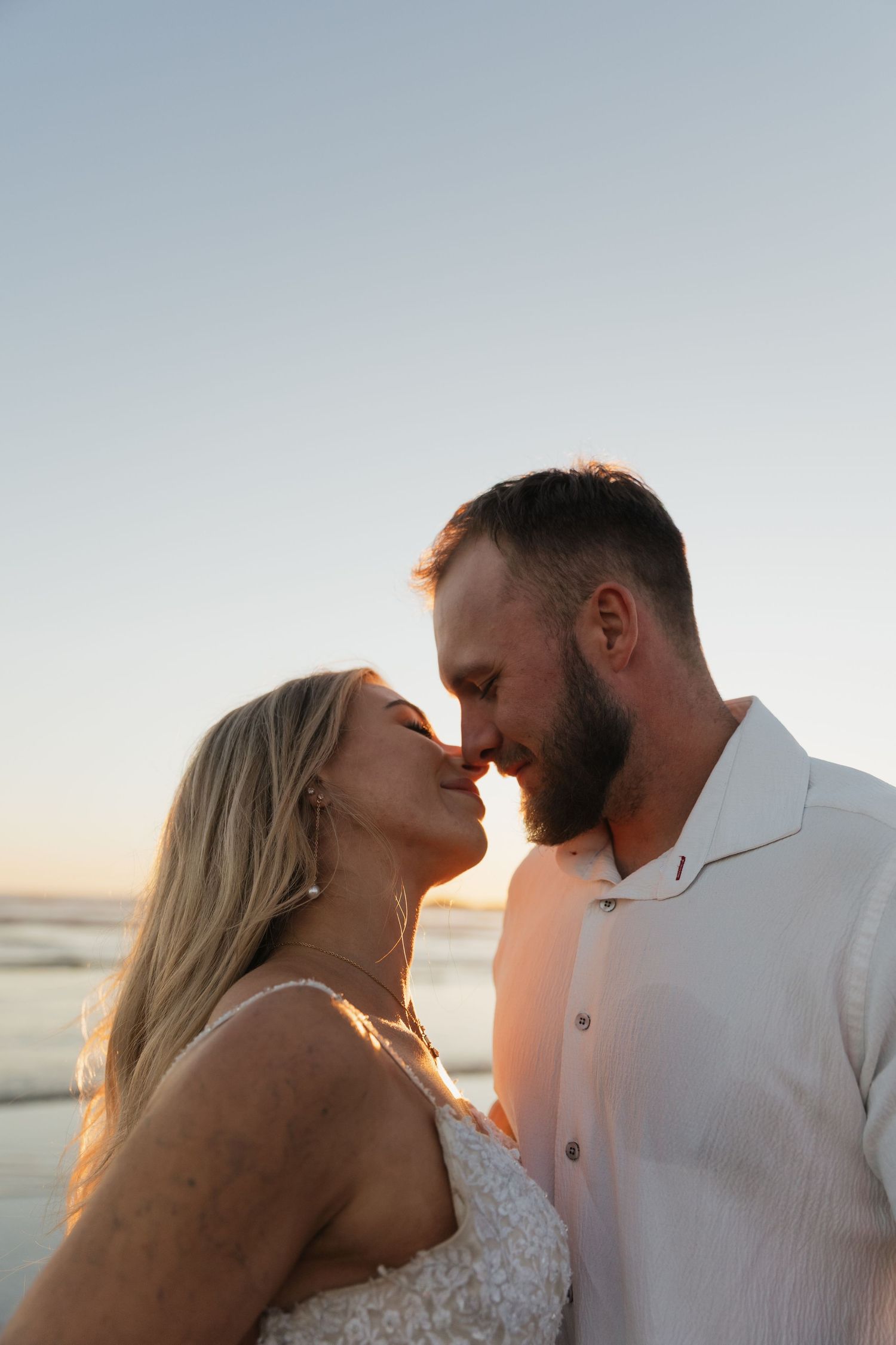 Couple sharing a romantic almost-kiss on the beach at sunset, wearing white wedding attire.