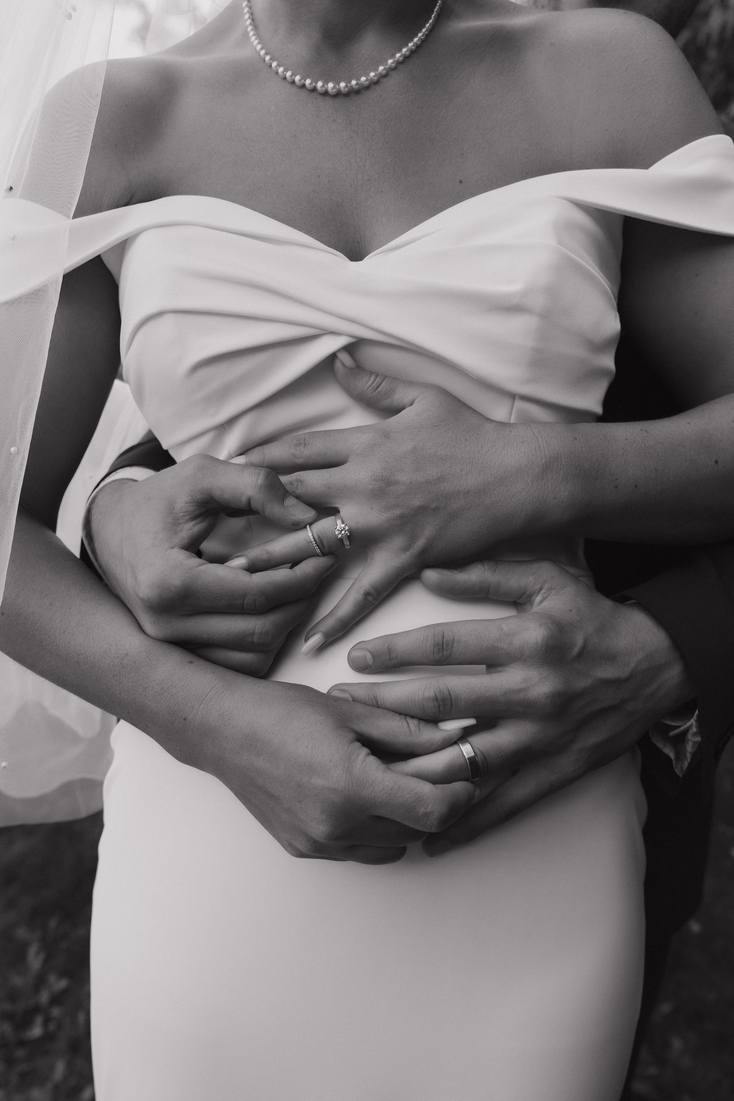 Black and white photo of groom embracing bride from behind, both wearing wedding rings.
