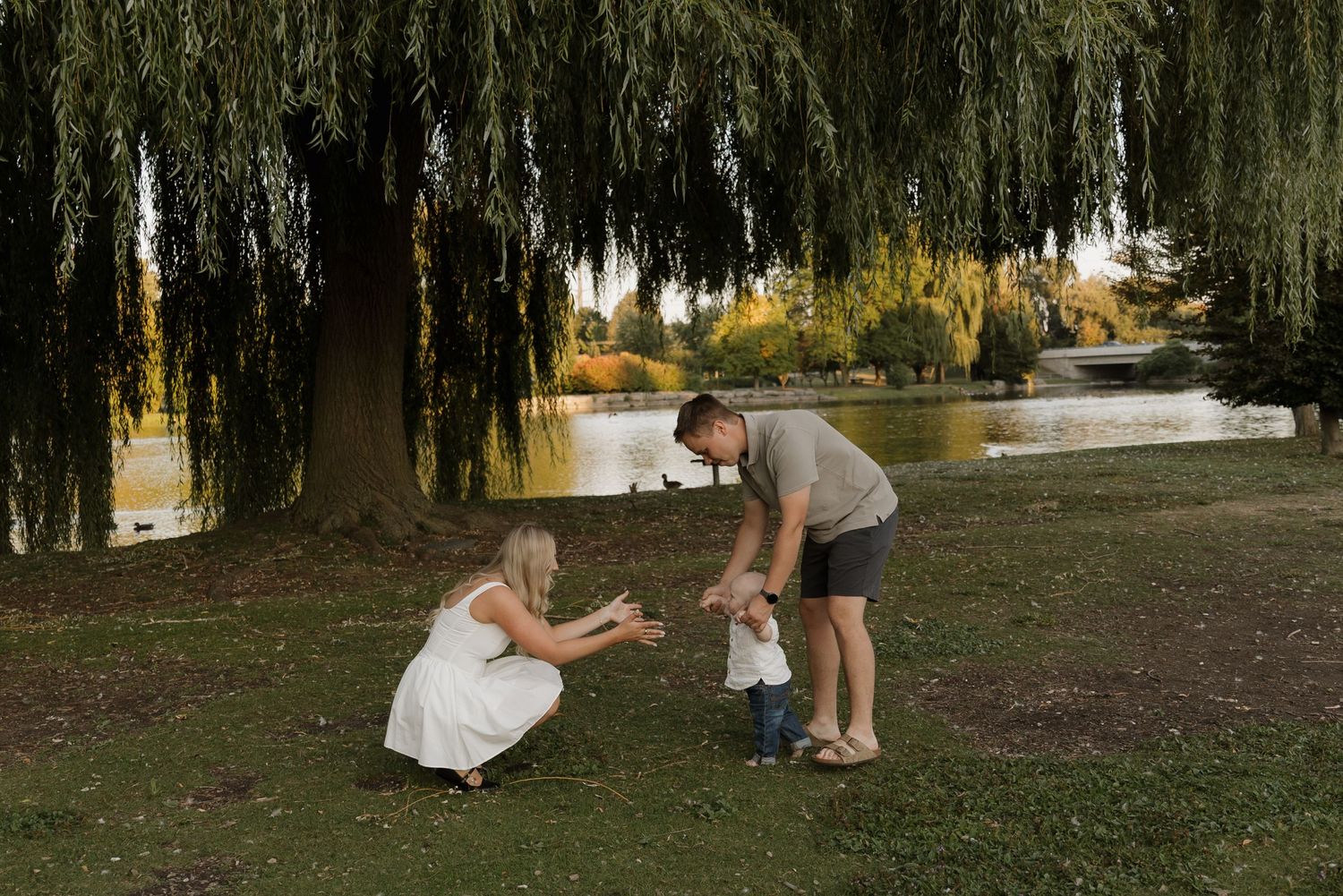 A young family with a toddler playing together under a weeping willow tree by a peaceful river.