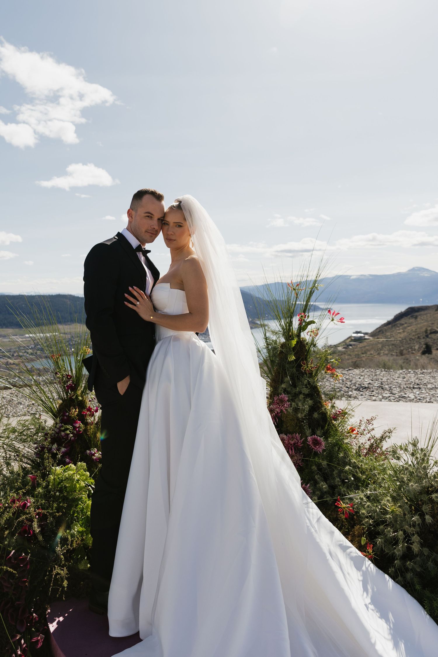 Bride and groom pose at outdoor wedding altar with mountain lake backdrop and lush floral arrangements.