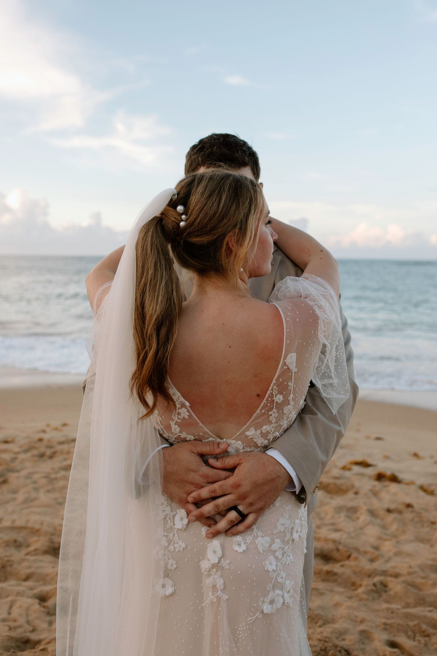 Bride and groom embrace facing the ocean on sandy beach at sunset wedding.