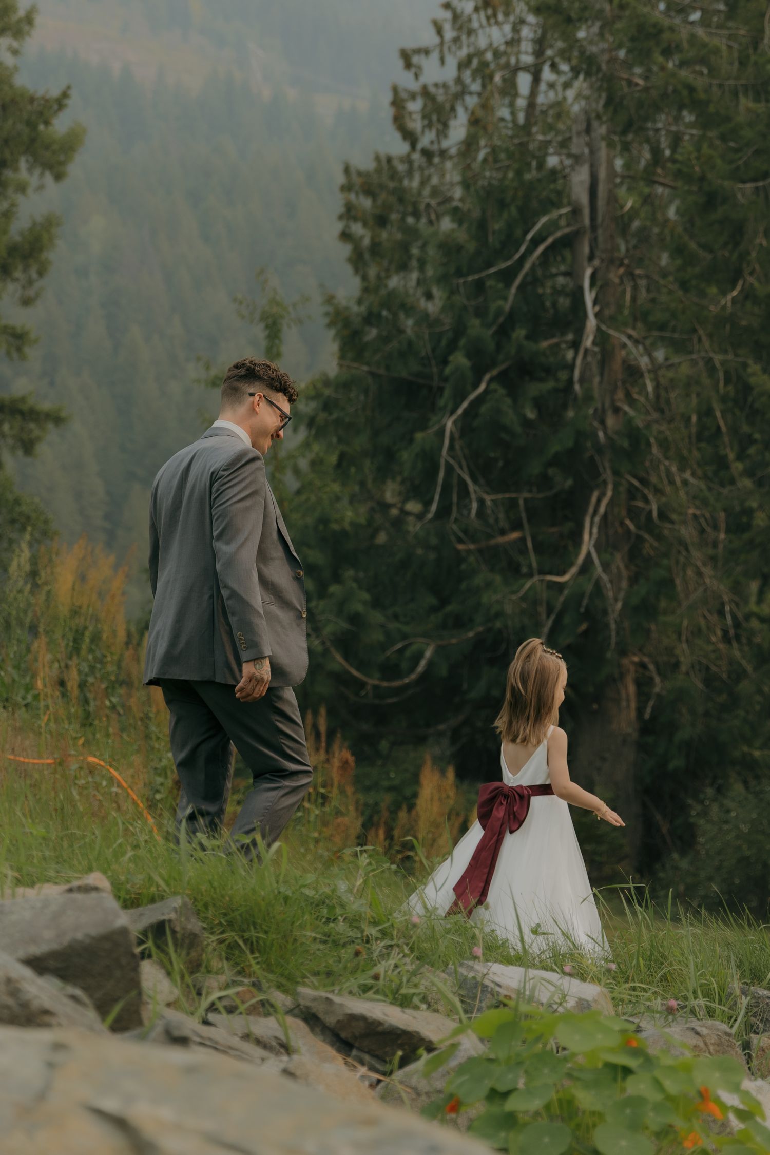 Man in gray suit walks with young flower girl in white dress with burgundy sash in forest.