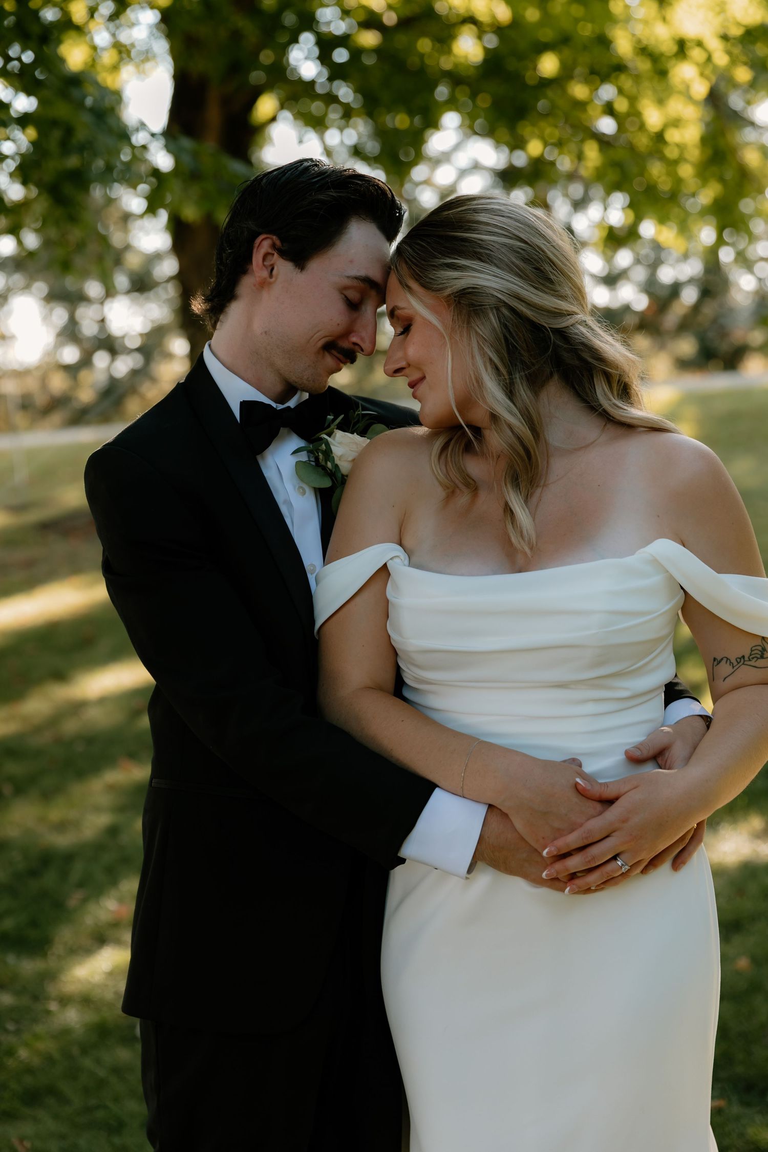 Bride and groom in black tuxedo sharing tender forehead touch embrace at golden hour outdoor wedding.