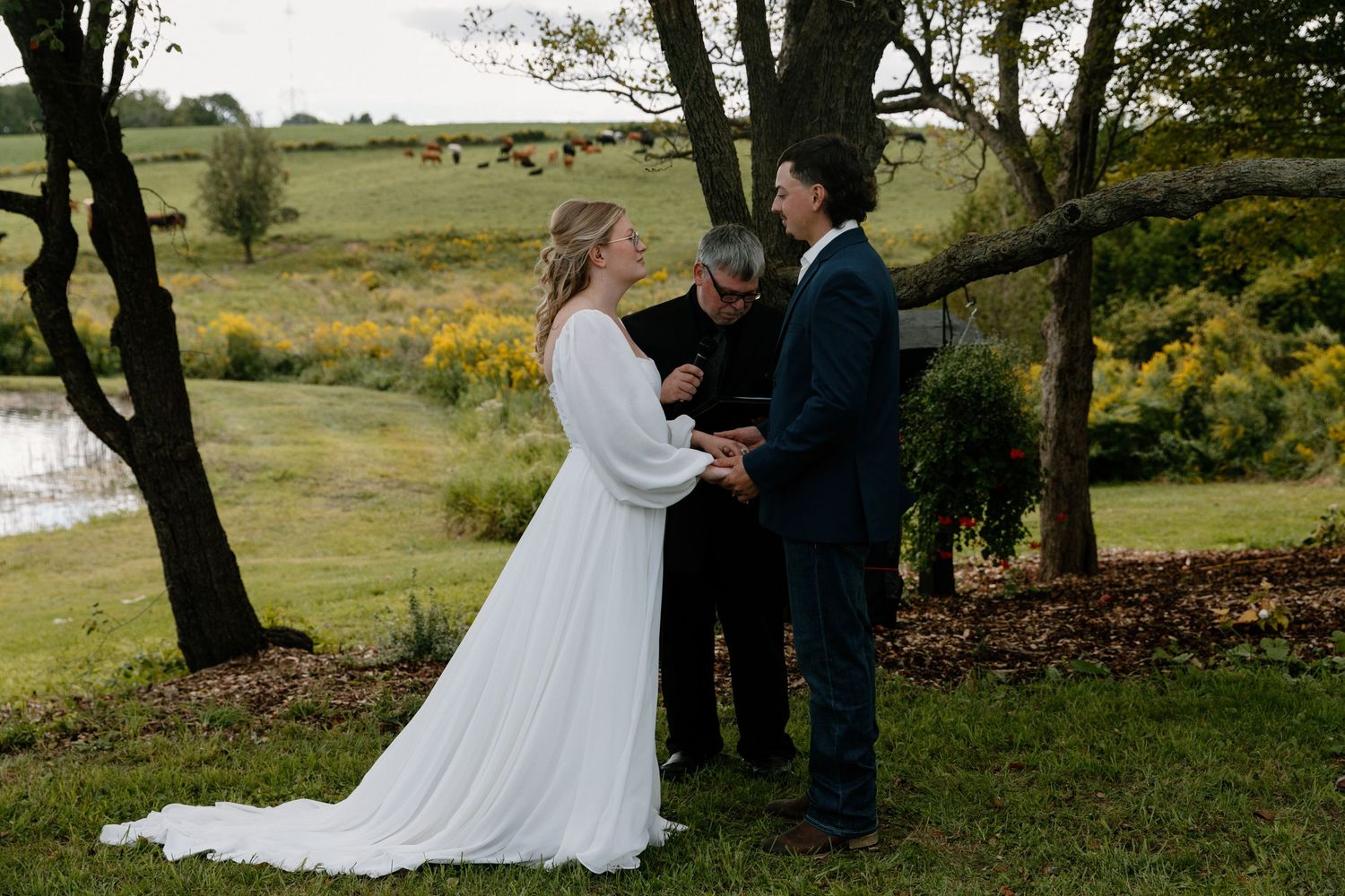 Bride and groom exchange vows outdoors with scenic pond and autumn foliage in background.