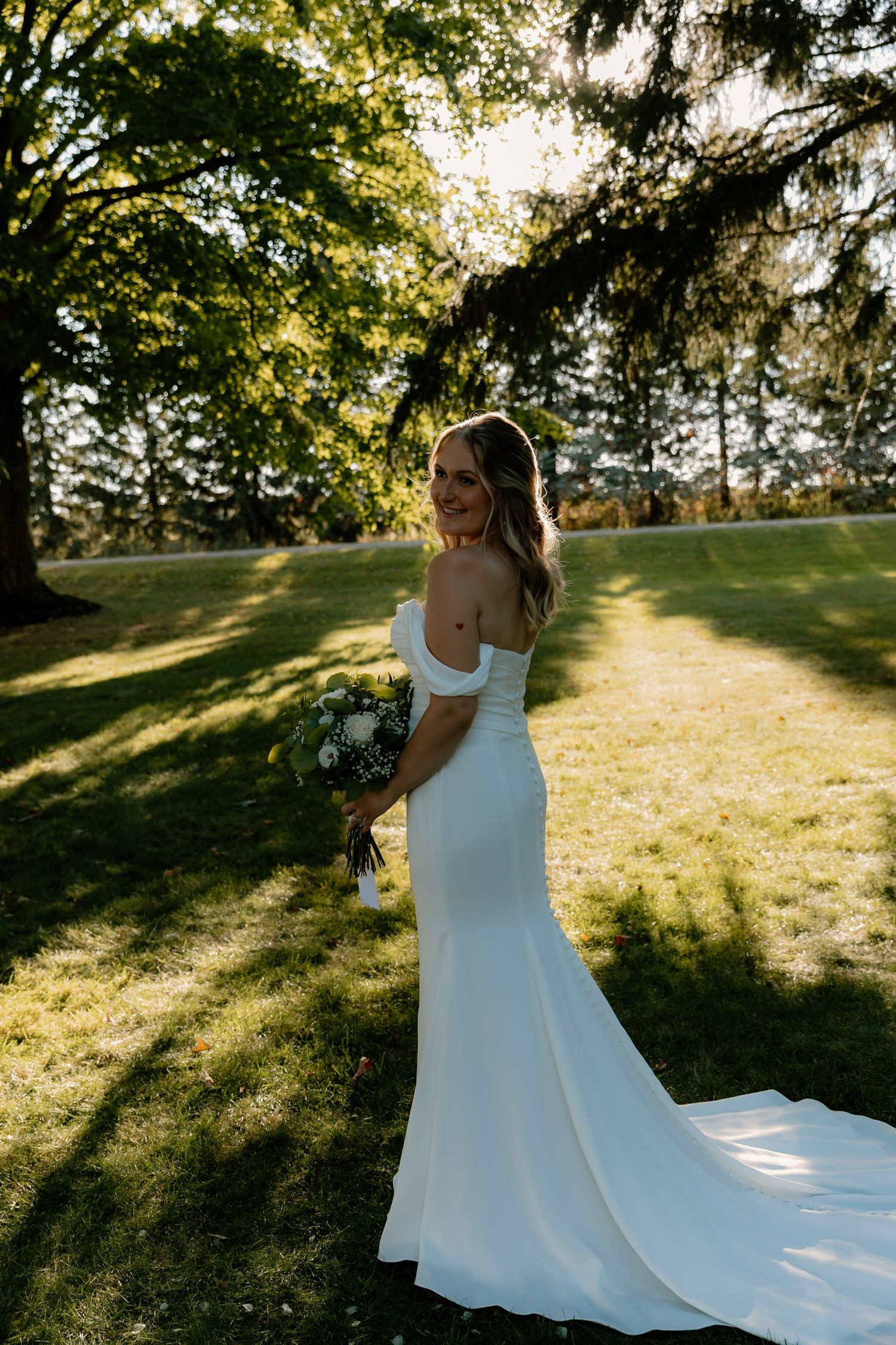 Elegant bride in off-shoulder white gown holding bouquet, smiling over shoulder in golden sunlit garden.