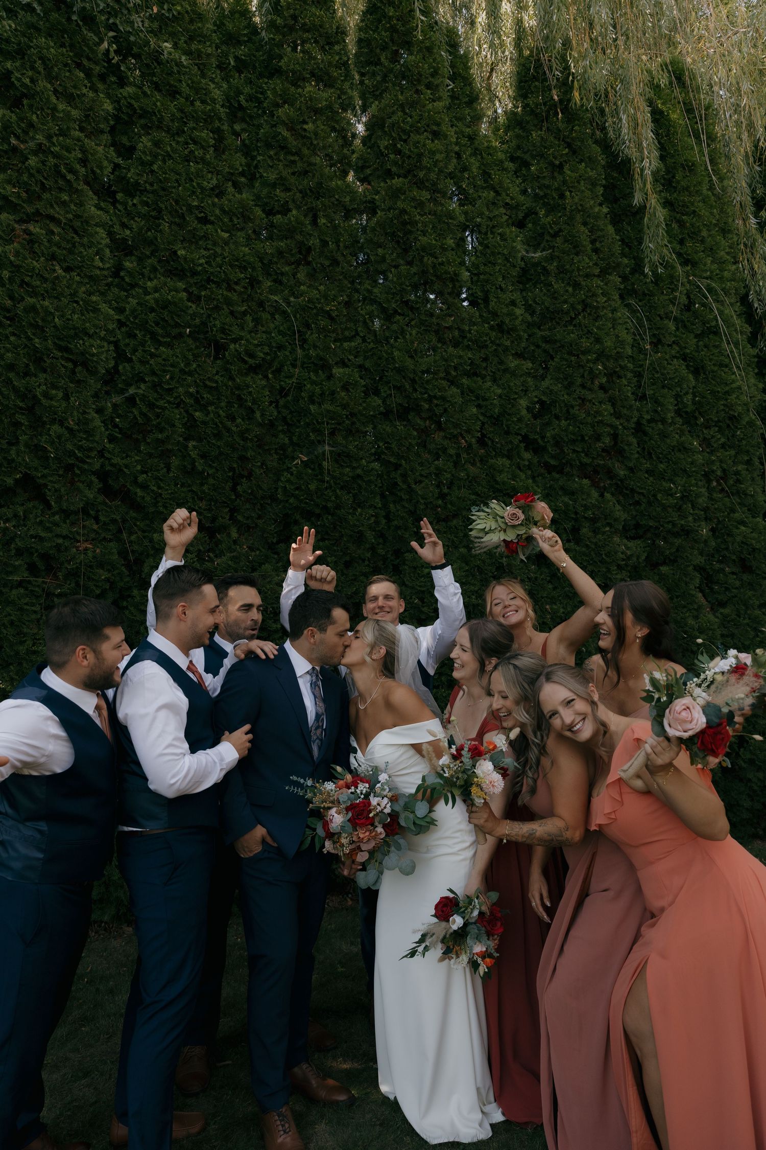 Joyful wedding party celebrates with raised arms before lush green hedge, bridesmaids in coral dresses, groomsmen in navy.