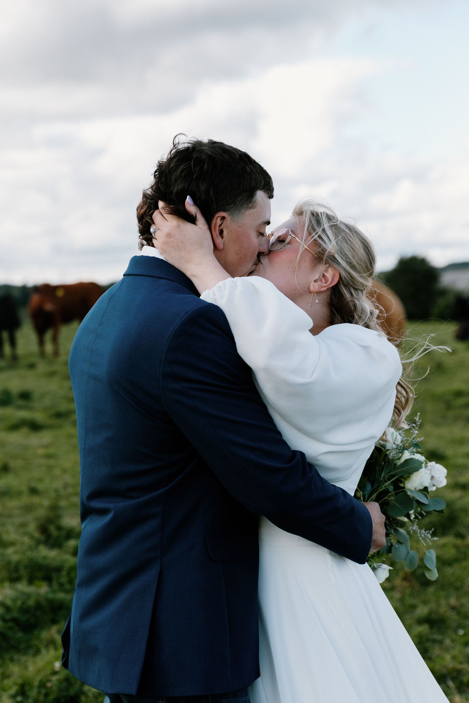 Newlyweds share a passionate kiss in a green field with cattle grazing behind them.