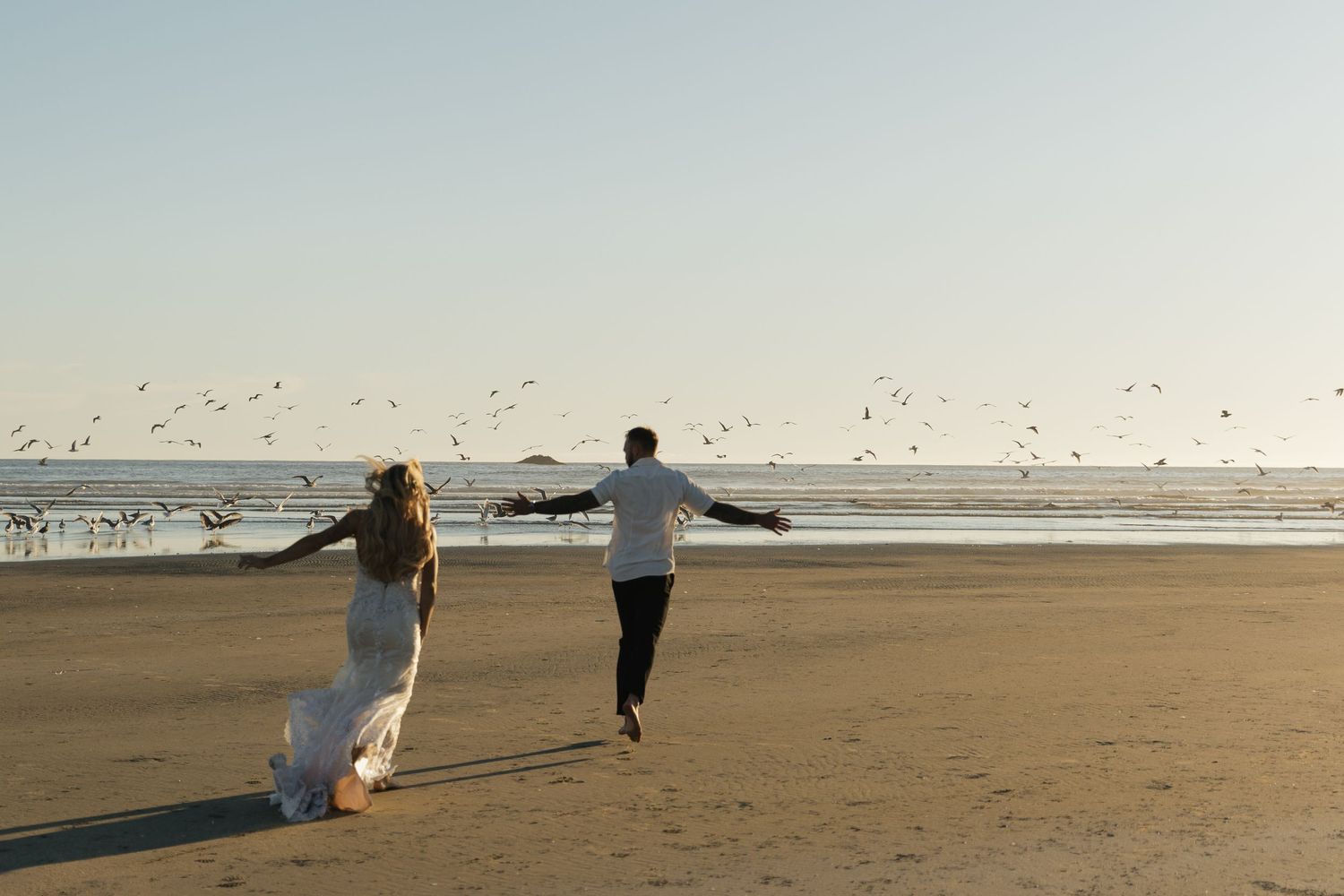 Bride and groom running on a sandy beach with arms outstretched, seagulls flying over the ocean at sunset.